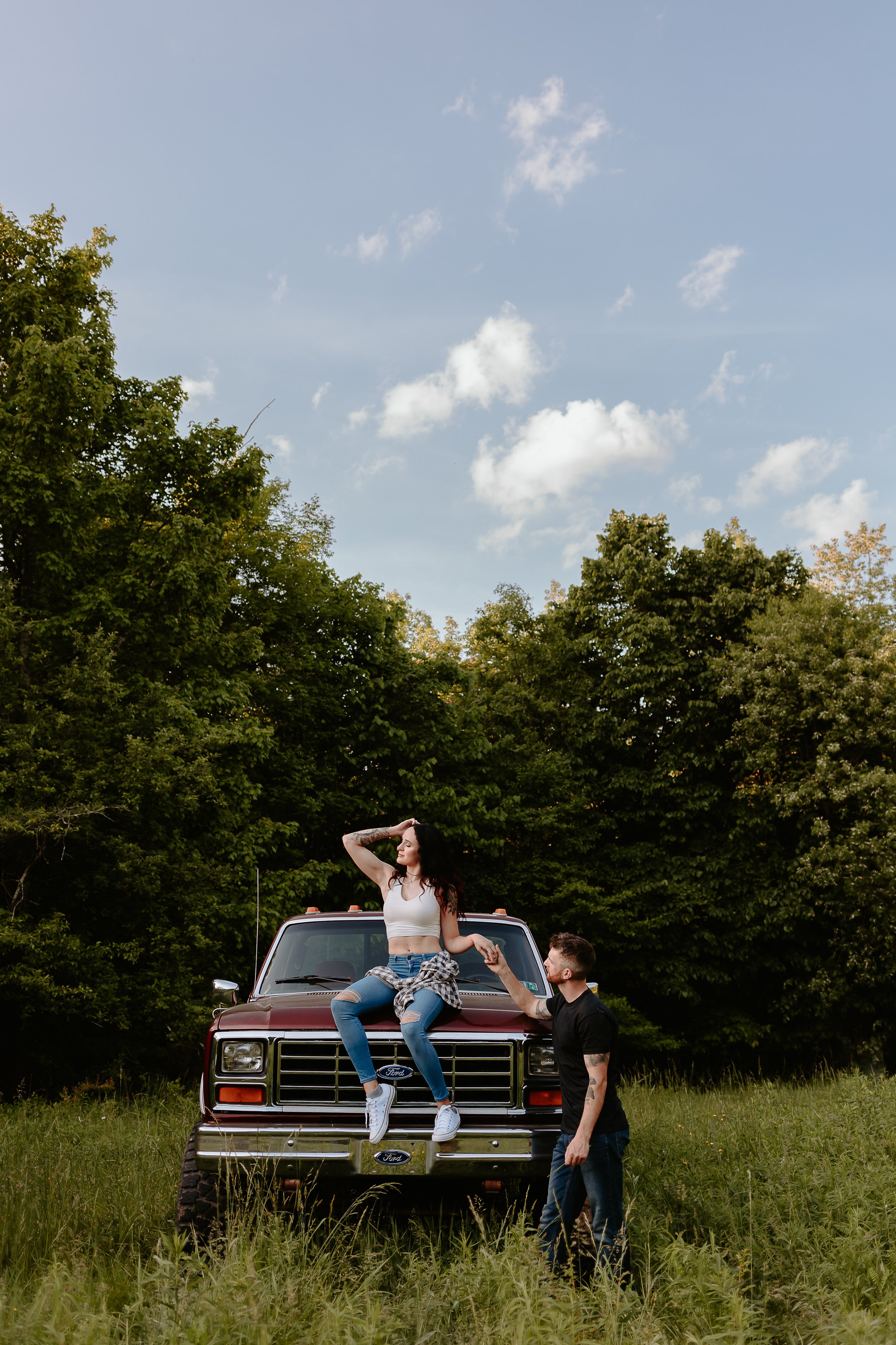 Woman sits on hood truck while fiancé holds her hand.