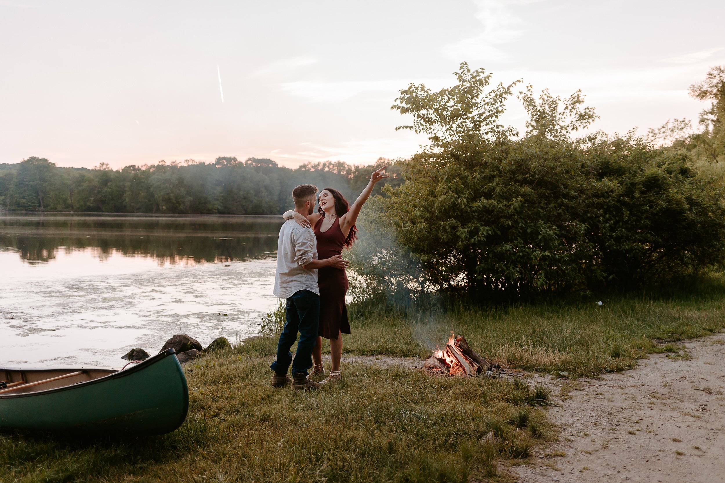 Woman and her fiancé dance by a campfire in front of a lake during sunset.