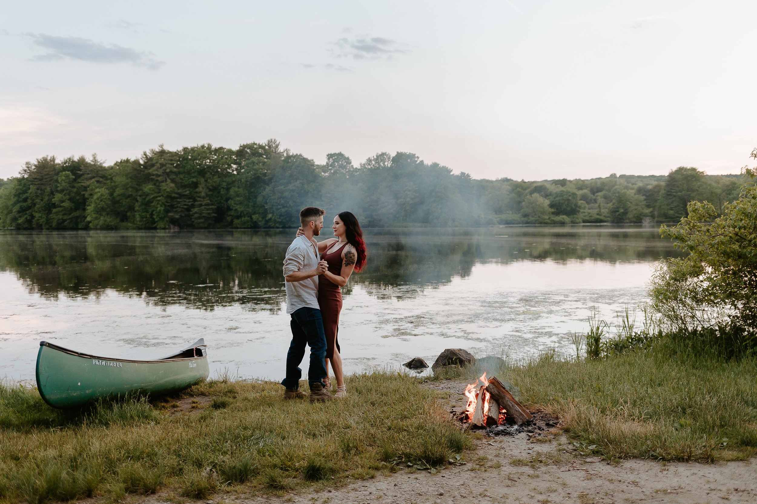 Man and fiancé dance by a campfire in front of a lake.