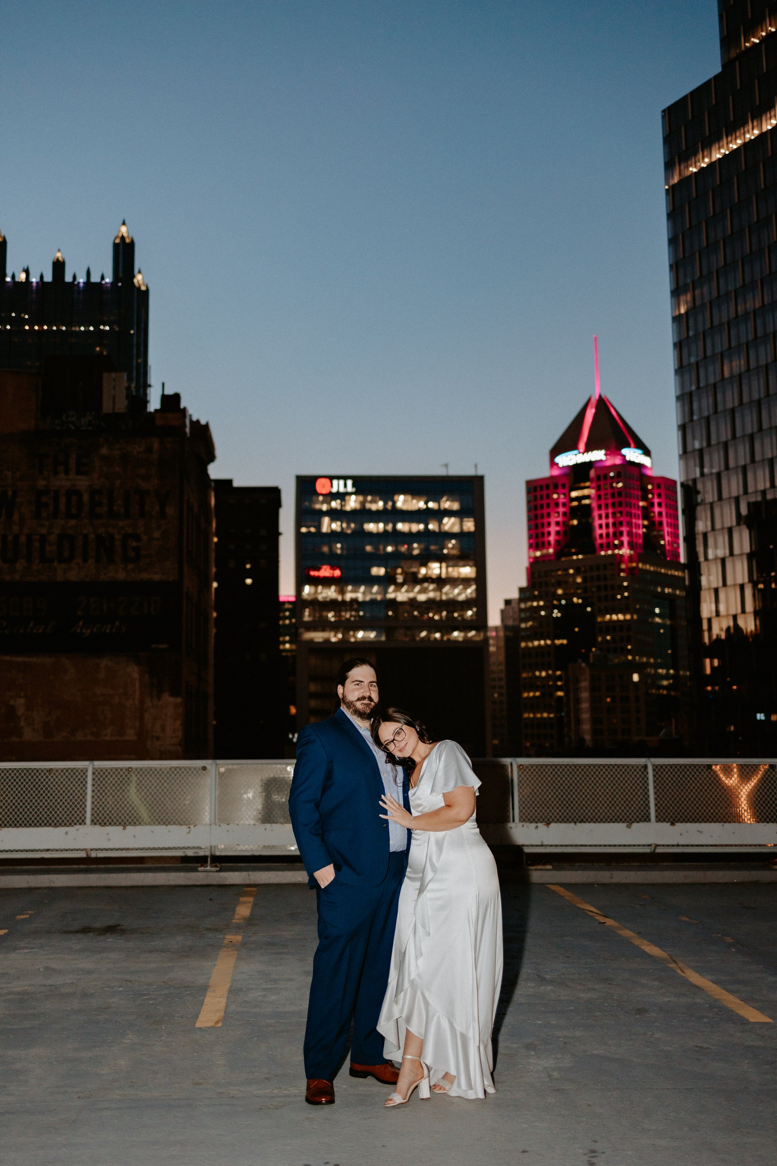 Woman and man embrace on parking garage roof with night time city skyline.