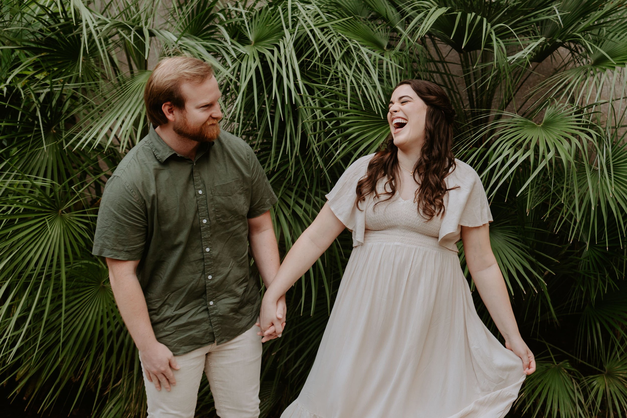 Man and woman hold hands laughing in front of green palm fronds.