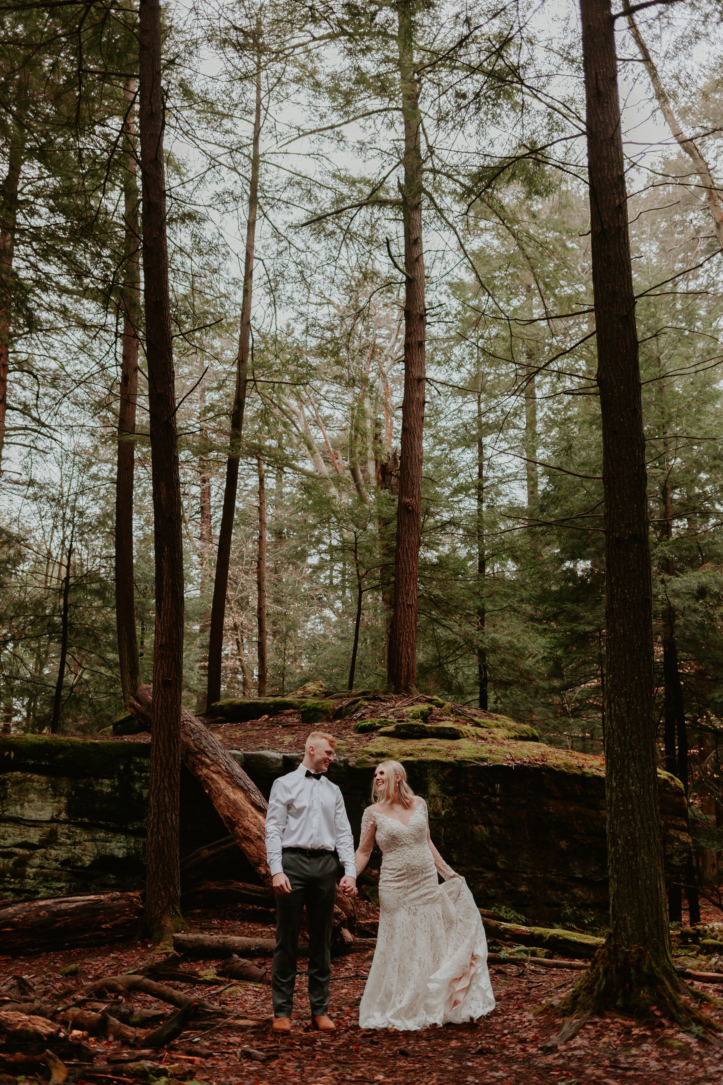 Groom holds bride's hand and looks at her while standing in front of large rock in the forest.