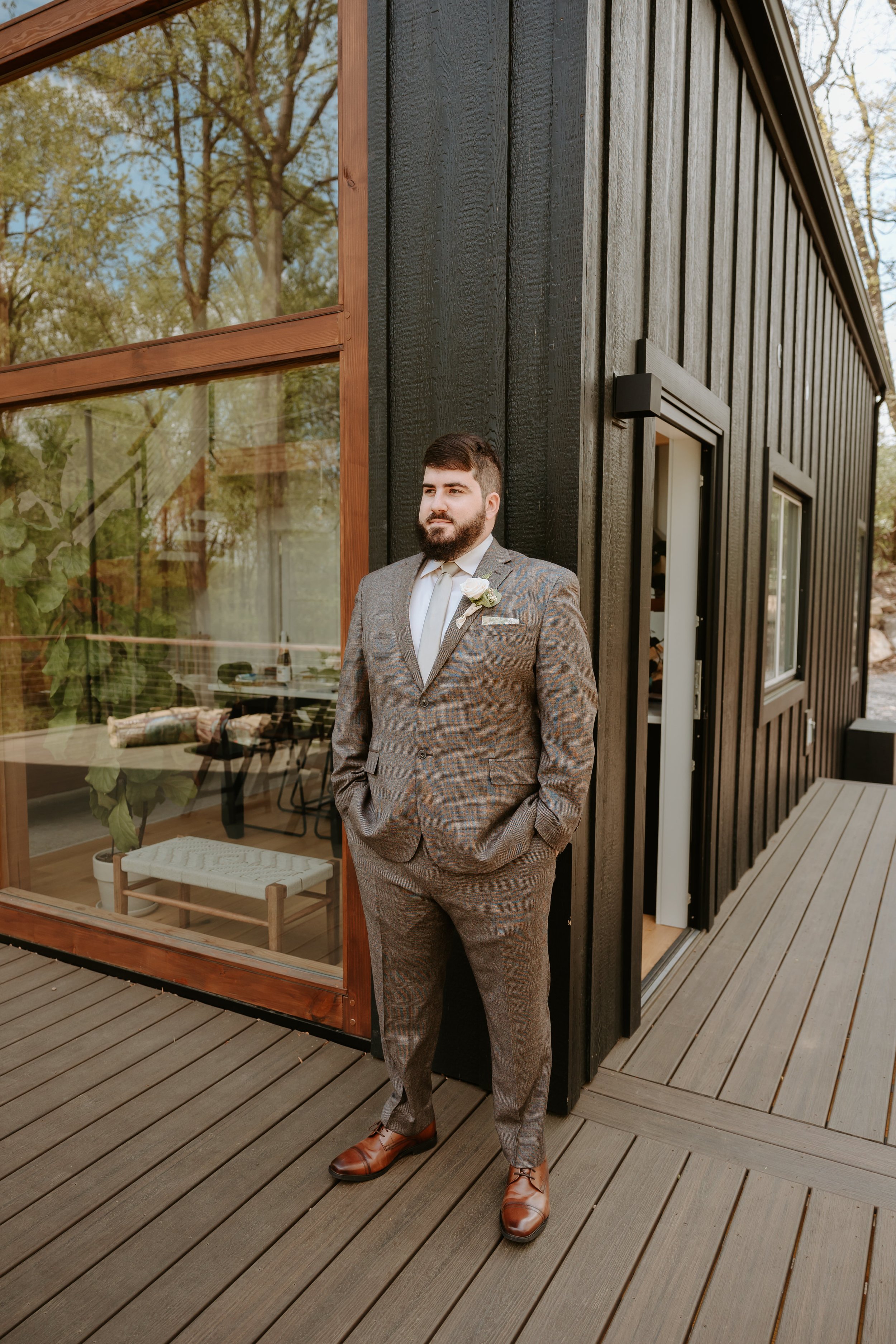 Man stands in front of black cabin while wearing a brown suit.