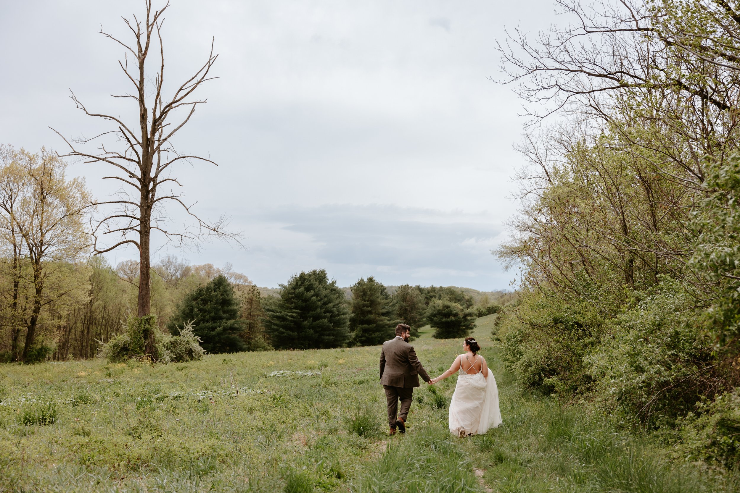Groom and bride walk holding hands in open green field. 