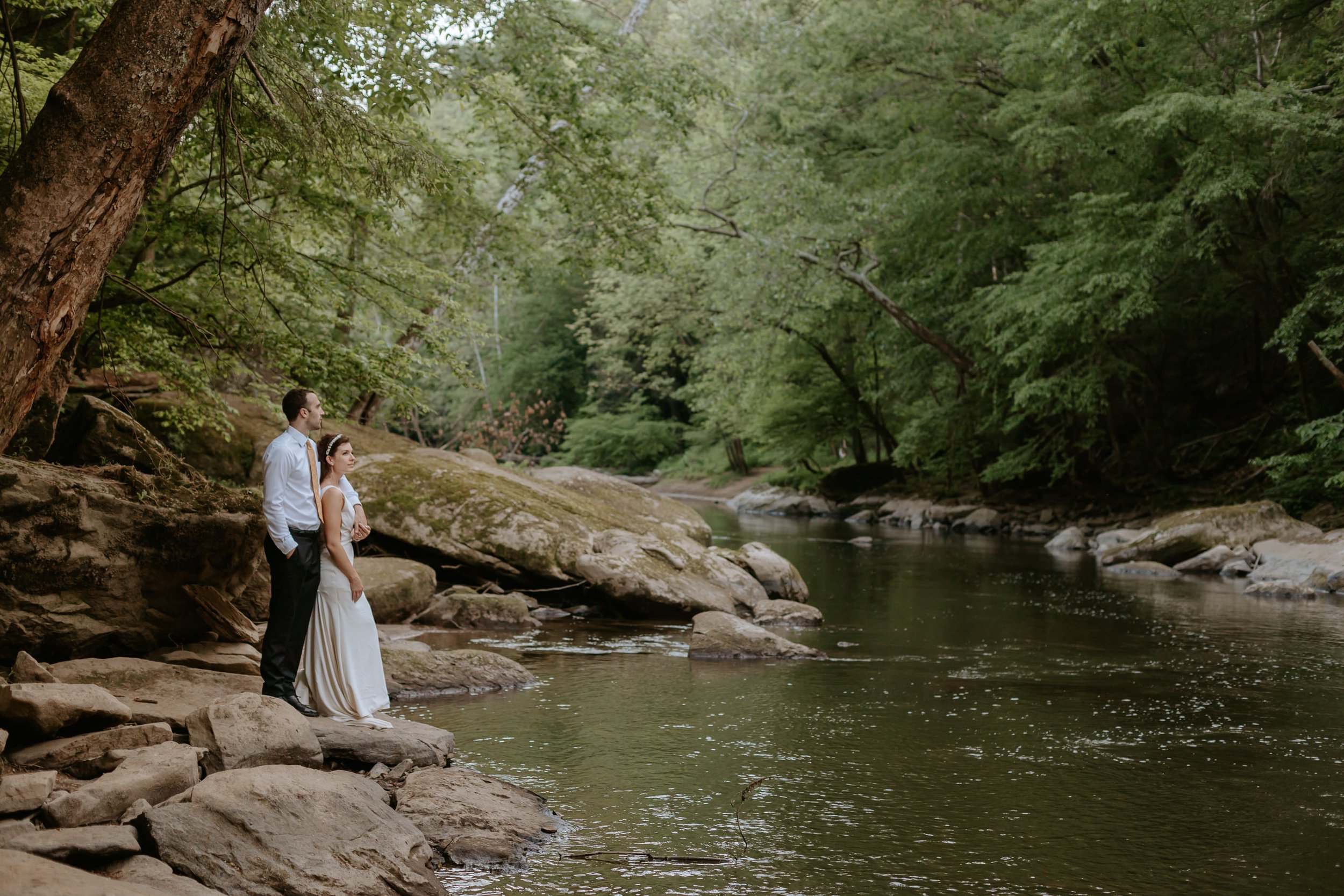 Couple stands together on rock looking at a creek.