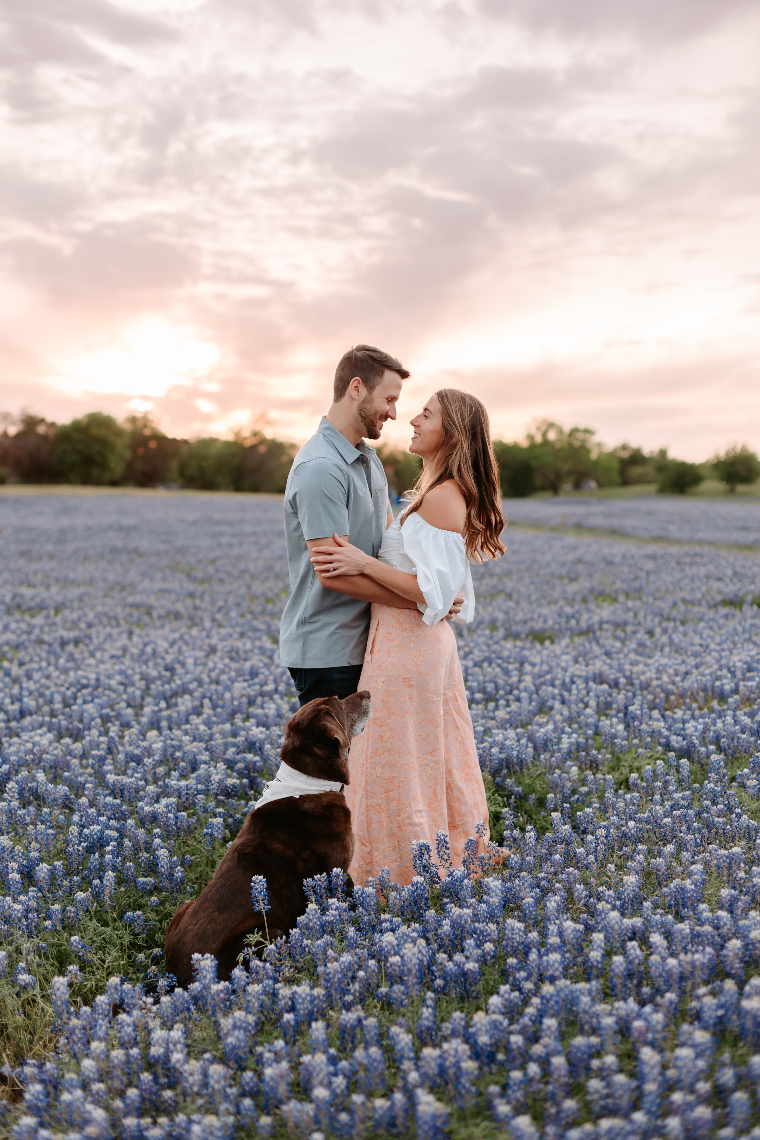 Man and woman embrace in a field of flowers with their dog.
