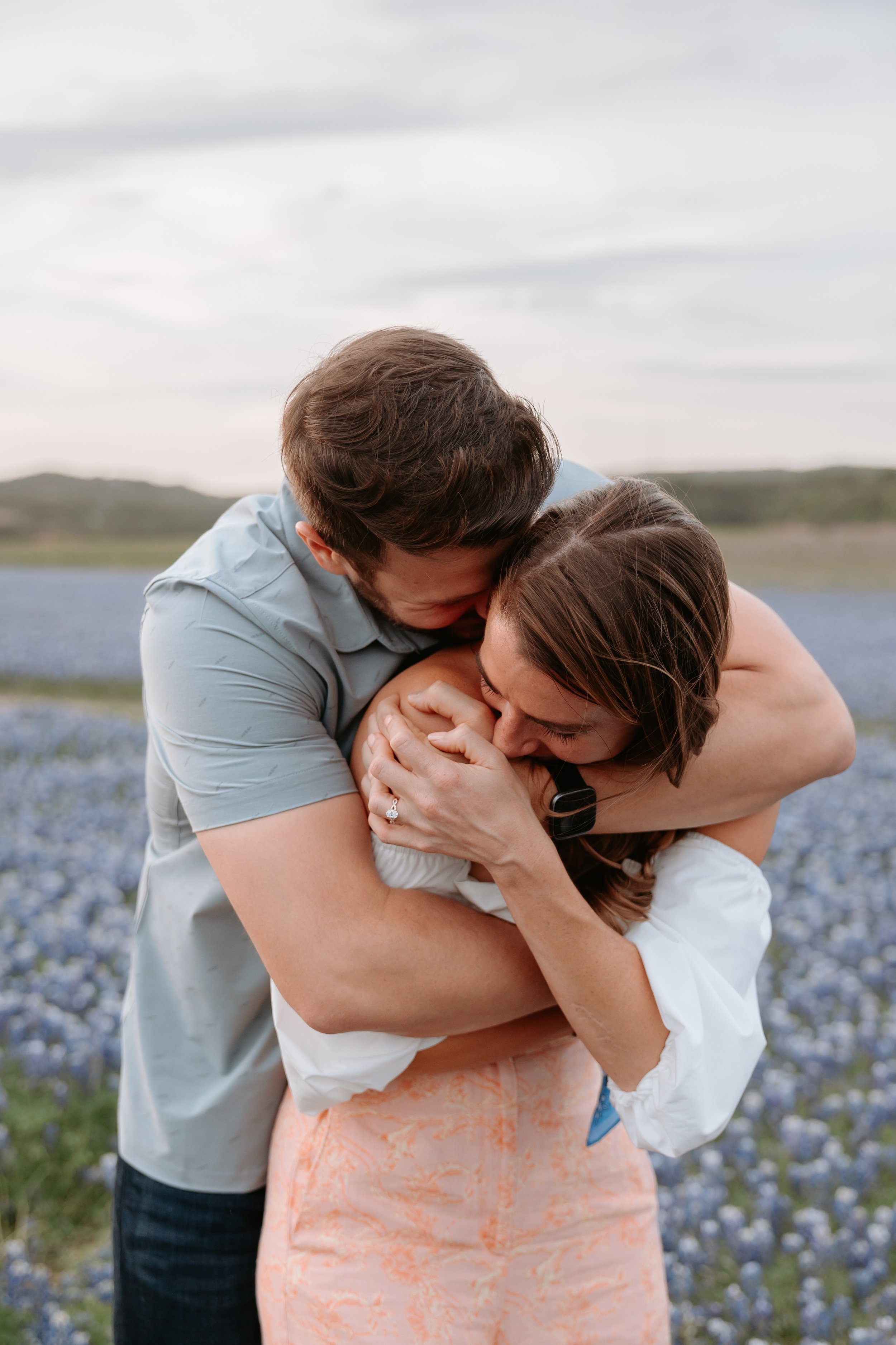 Man hugs woman from behind in a field of blue flowers.