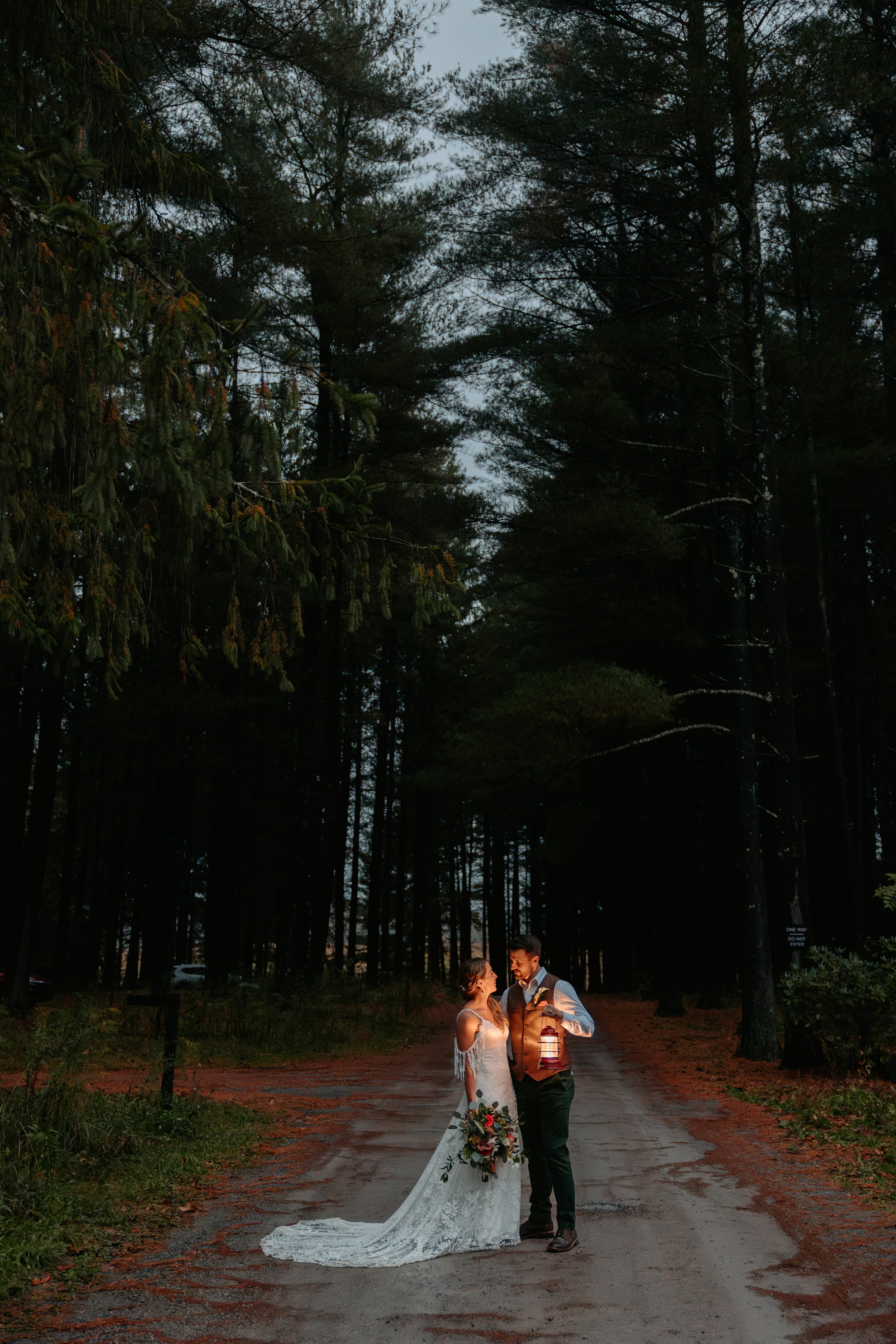 Bride and groom stand on gravel road with a lantern.