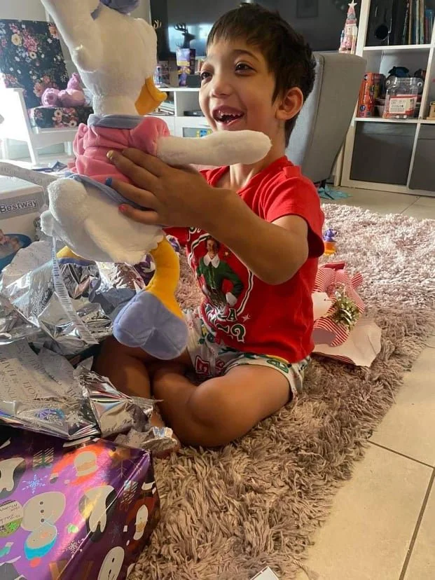 A little boy with Jansen's Disease opening up a  gift of a stuffed duck dressed in a pink dress, for Christmas