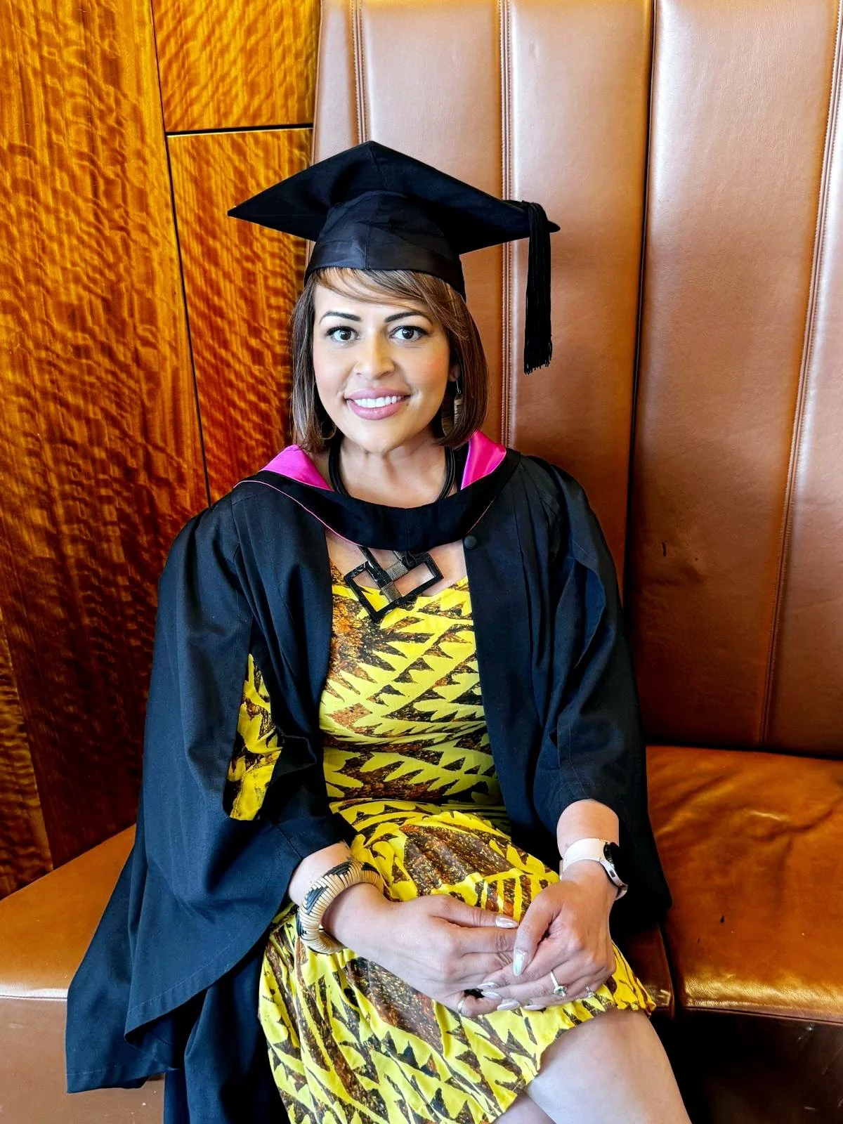 A woman dressed in a graduation gown and cap, sitting on a leather bench, smiling at the camera.