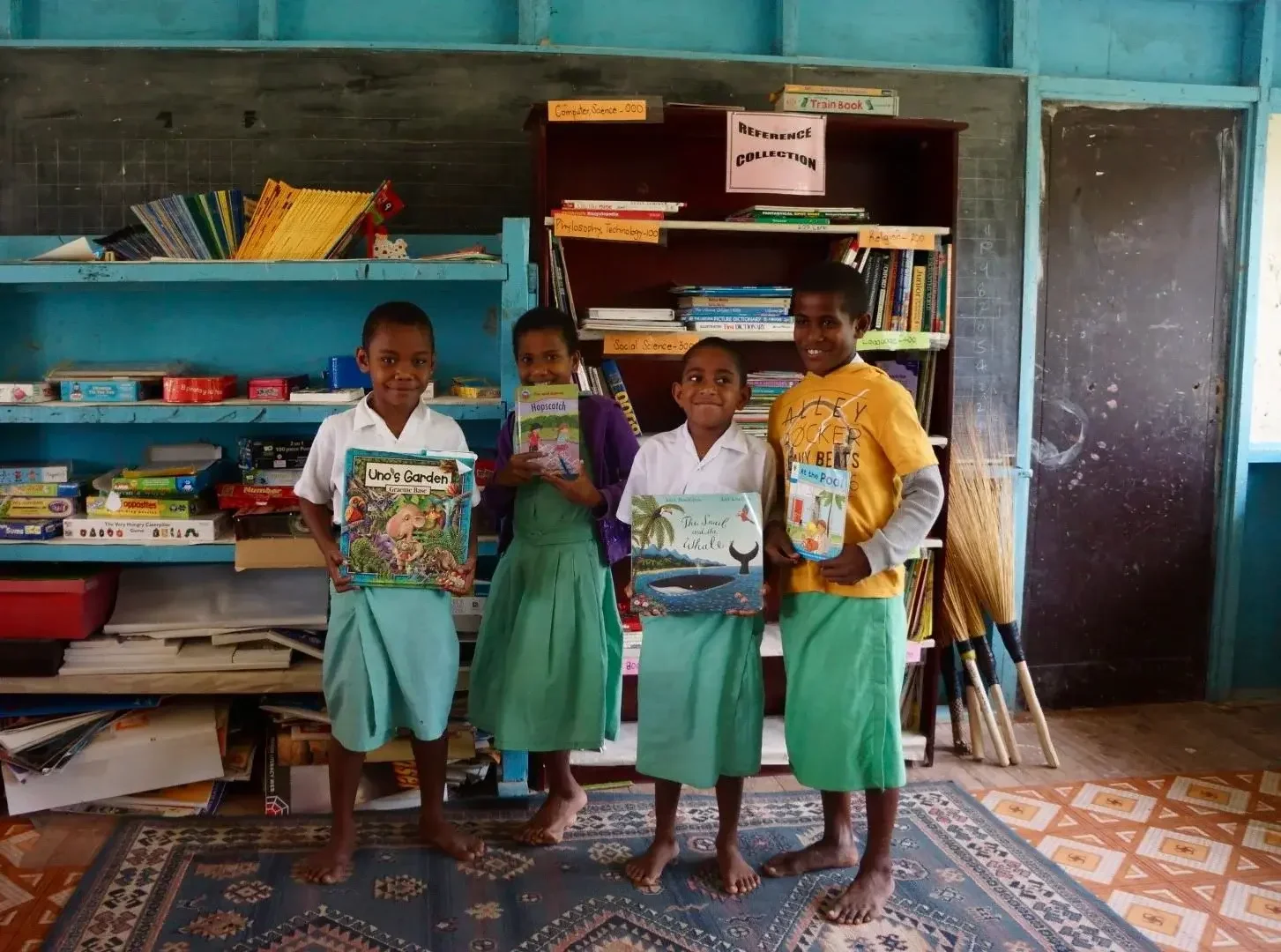 Four Fijian children holding books with bookshelves behind them .
