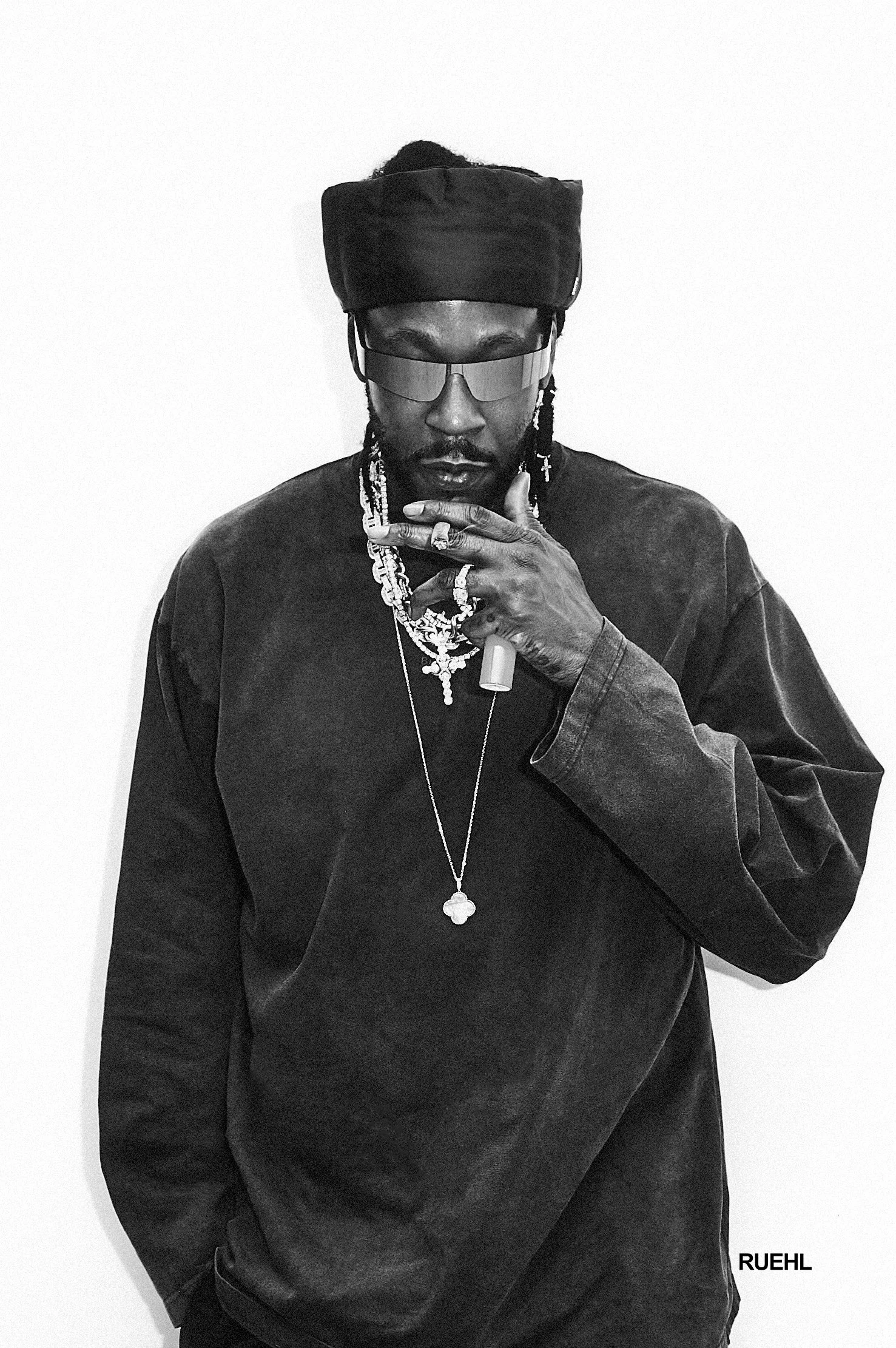 A man wearing a black headwrap, large sunglasses, and jewelry, including multiple necklaces and earrings, poses with his hand near his face against a plain white background.