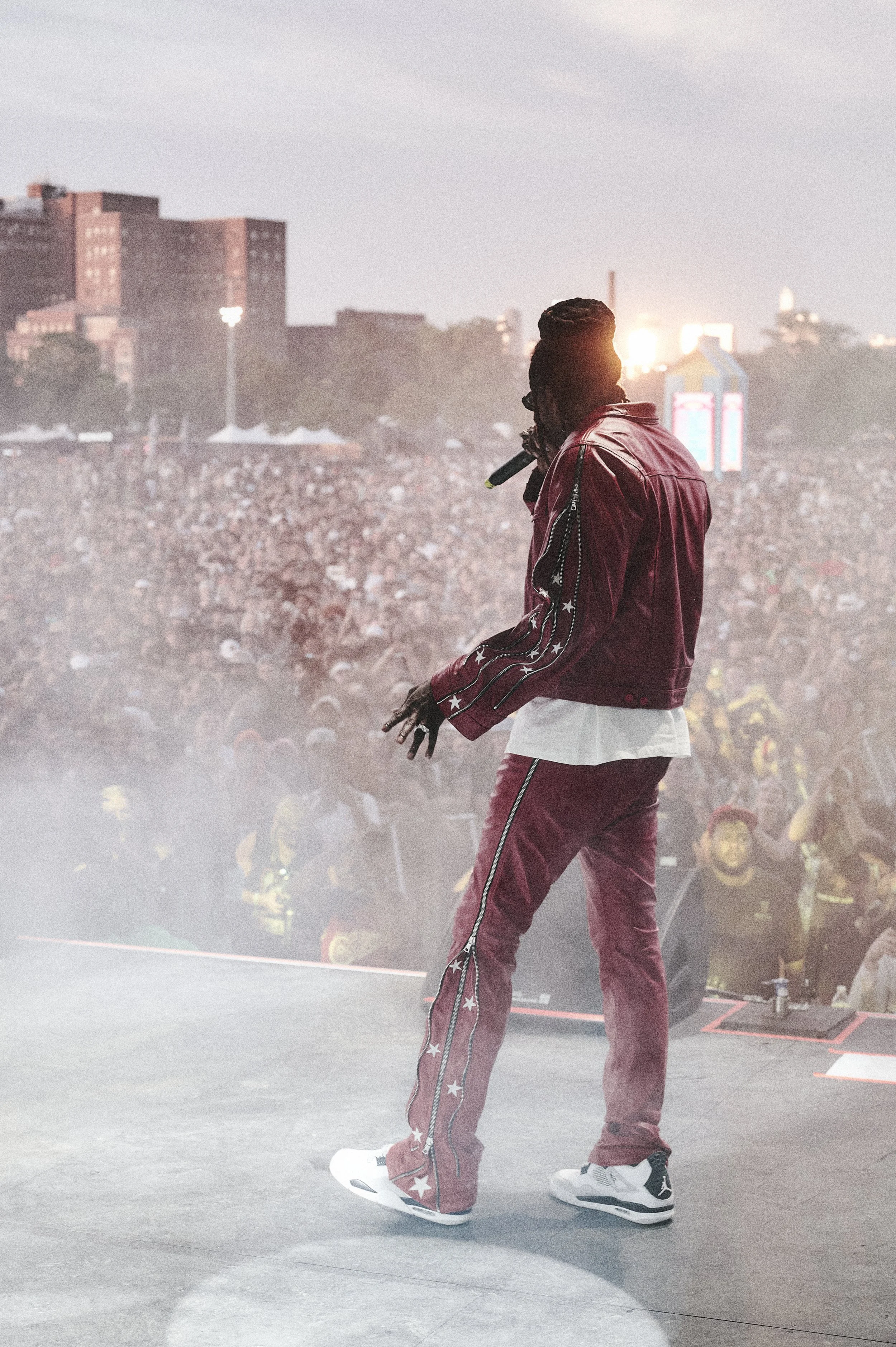 A performer with braids wearing a red jacket and matching pants stands on stage with a microphone, facing a large crowd at an outdoor concert during sunset.