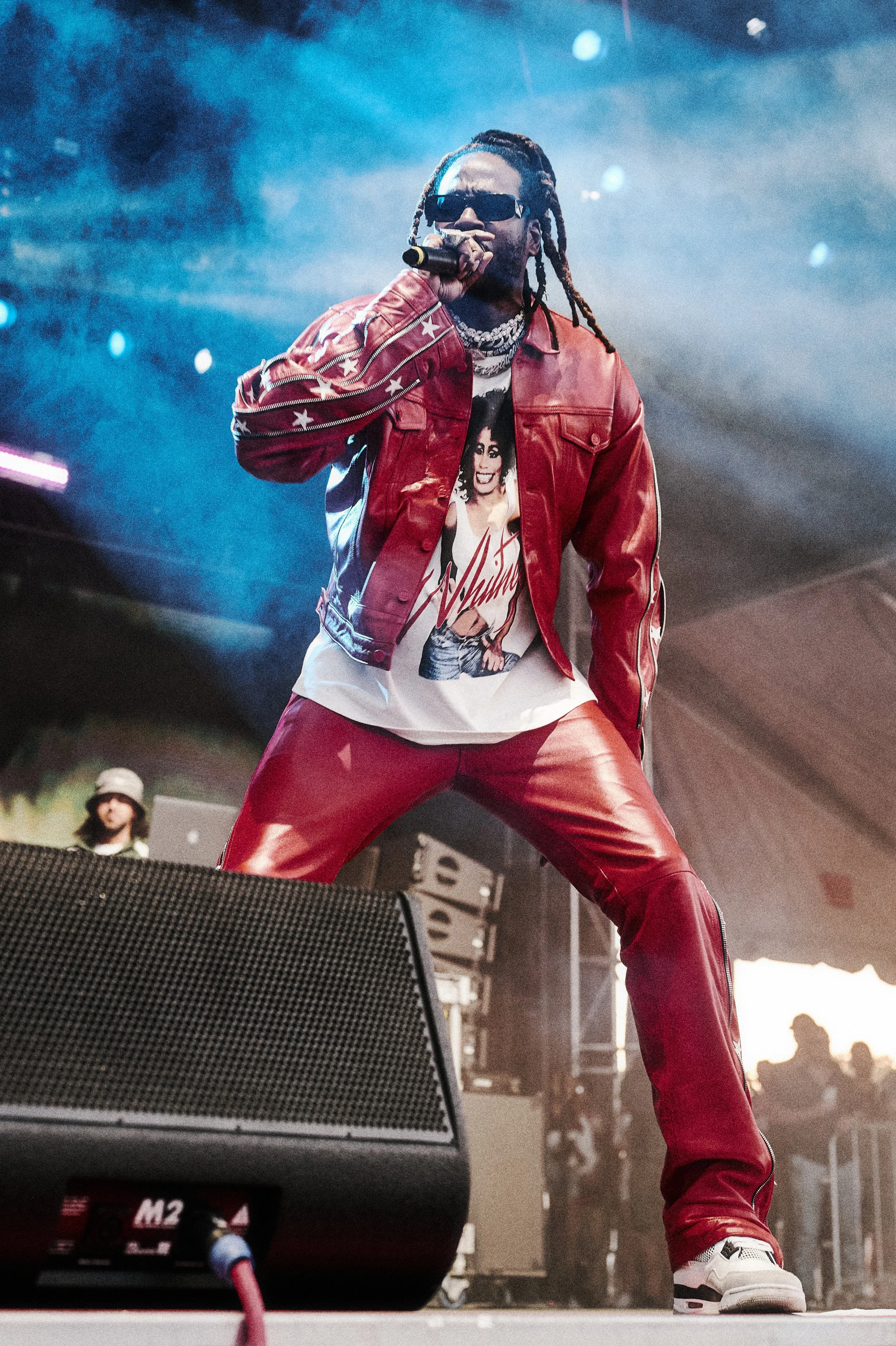 Male rapper performing on stage, wearing red leather jacket and pants, sunglasses, and a graphic t-shirt, holding a microphone.
