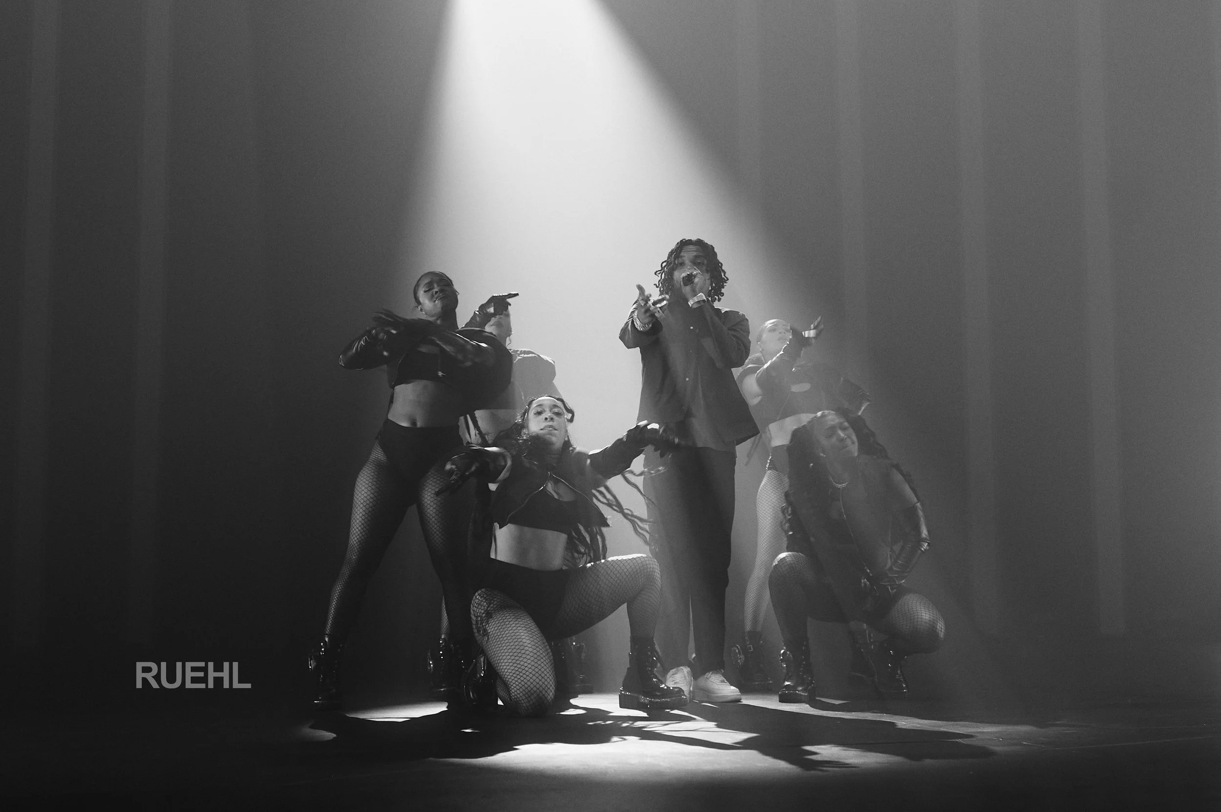 A black and white photo of a musical performance with one singer and four dancers on stage, illuminated by spotlight, with the word 'RUEHL' in the bottom left corner.