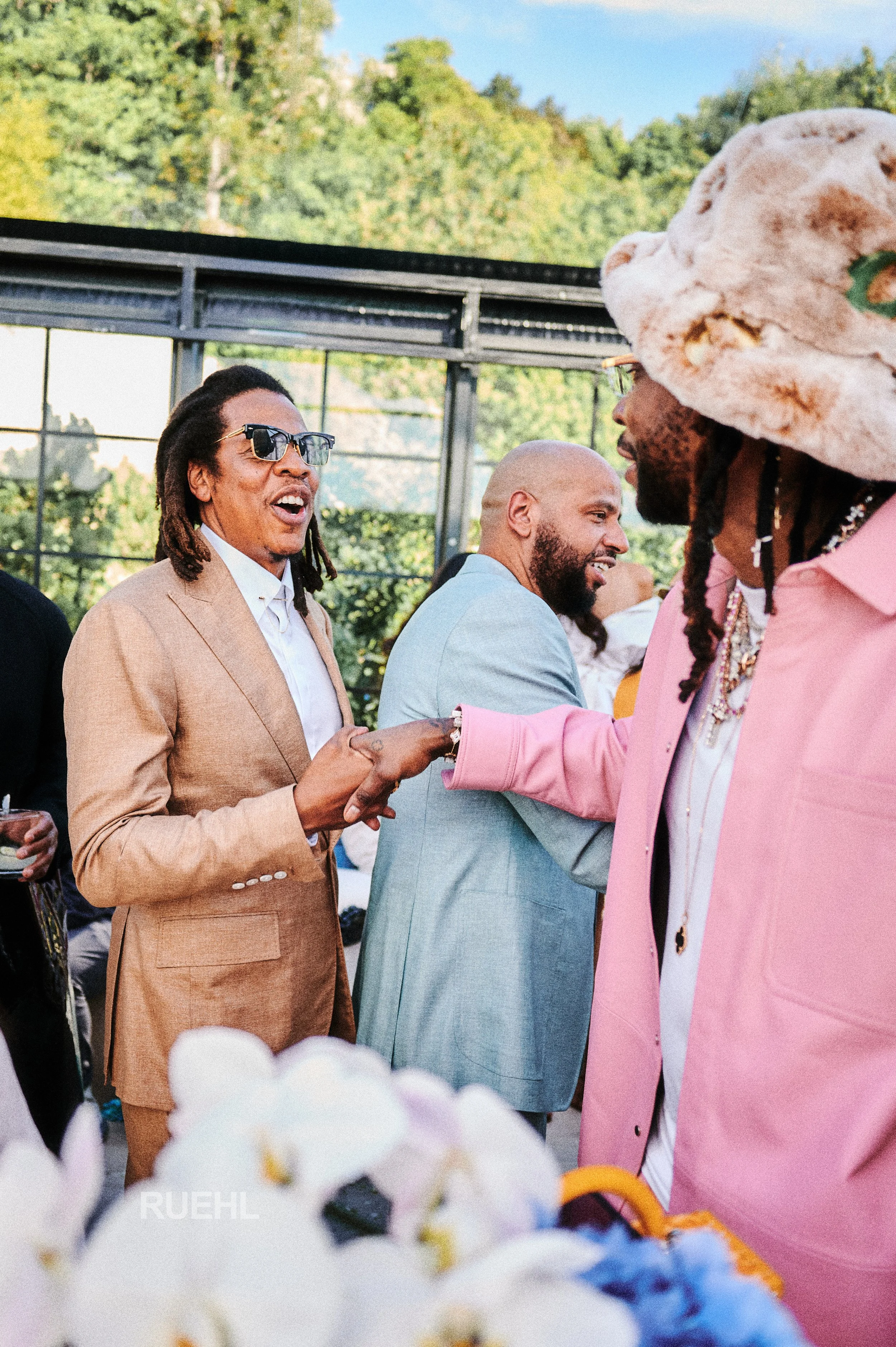 Three men are talking and smiling at an outdoor event with trees and a glass building in the background. One man wears sunglasses and a tan suit, another a light blue suit, and a third wears a pink suit with a large, furry hat.