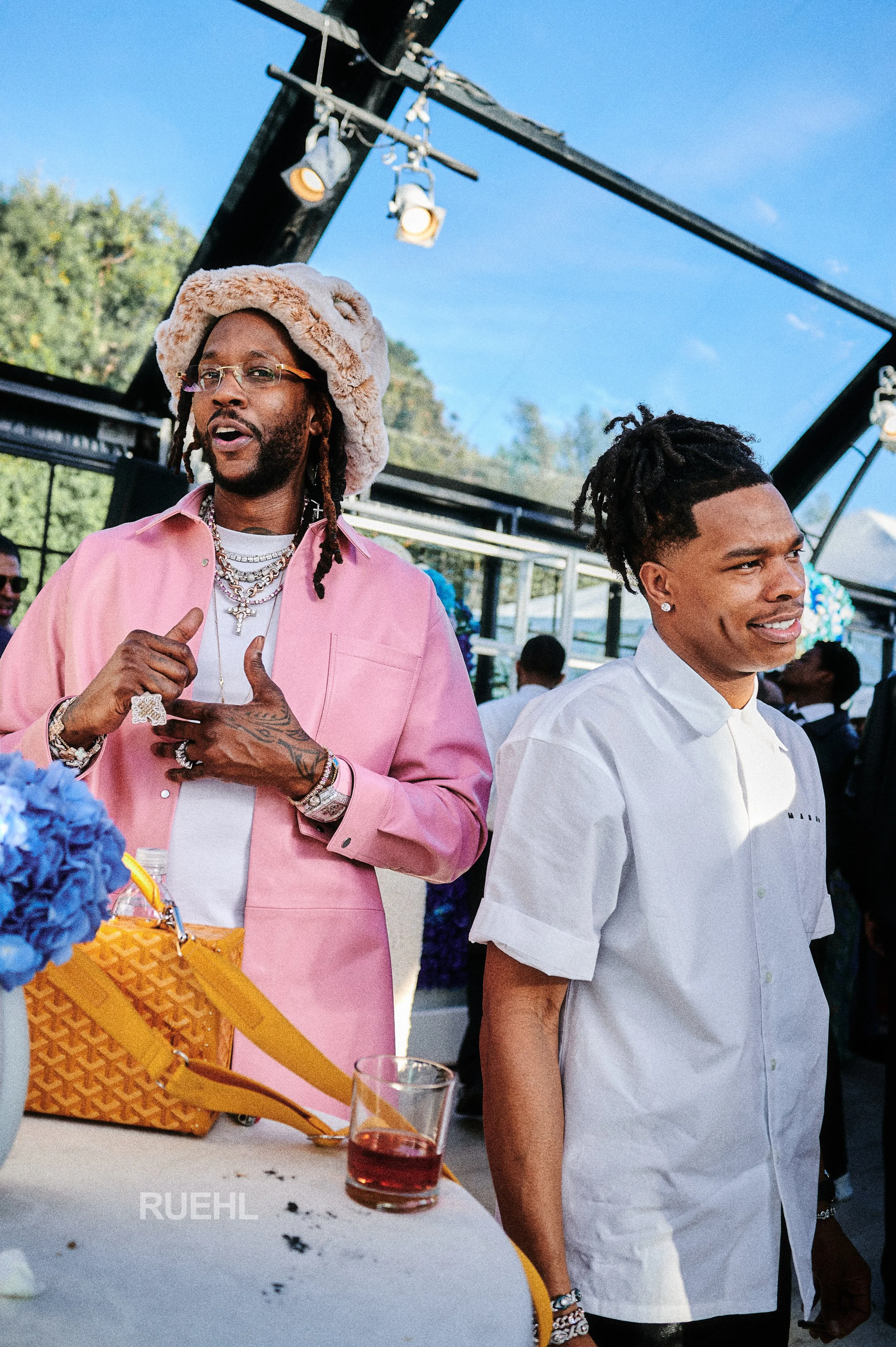 Two men at a social event, one wearing a pink jacket and fluffy hat, accessorized with jewelry, and the other in a white shirt with rolled-up sleeves and earrings, standing outdoors under a black geometric structure with a blue sky overhead.