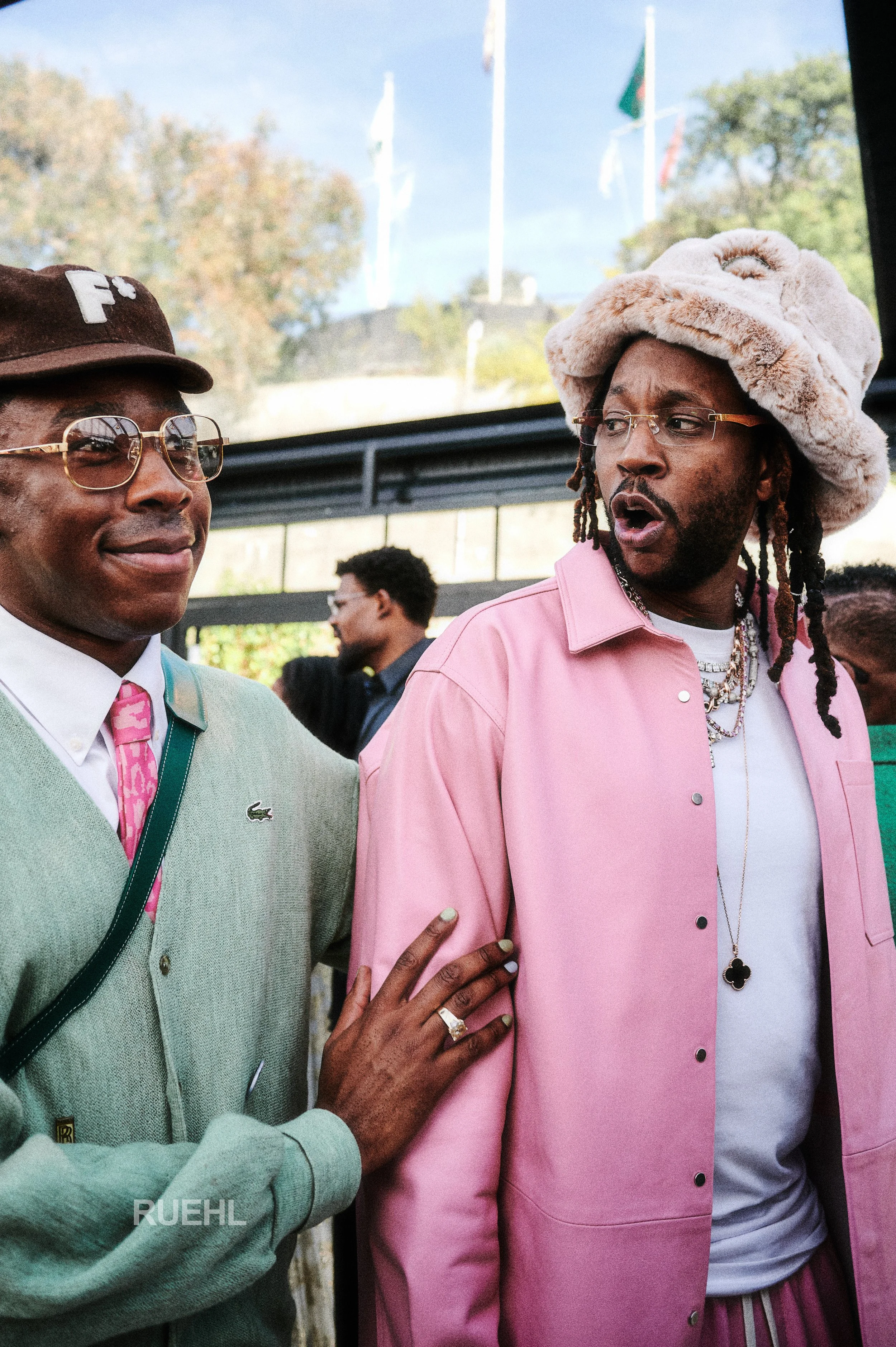 Two men in colorful clothing and glasses, one with a pink hat, engaged in conversation at an outdoor event with flags and trees in the background.