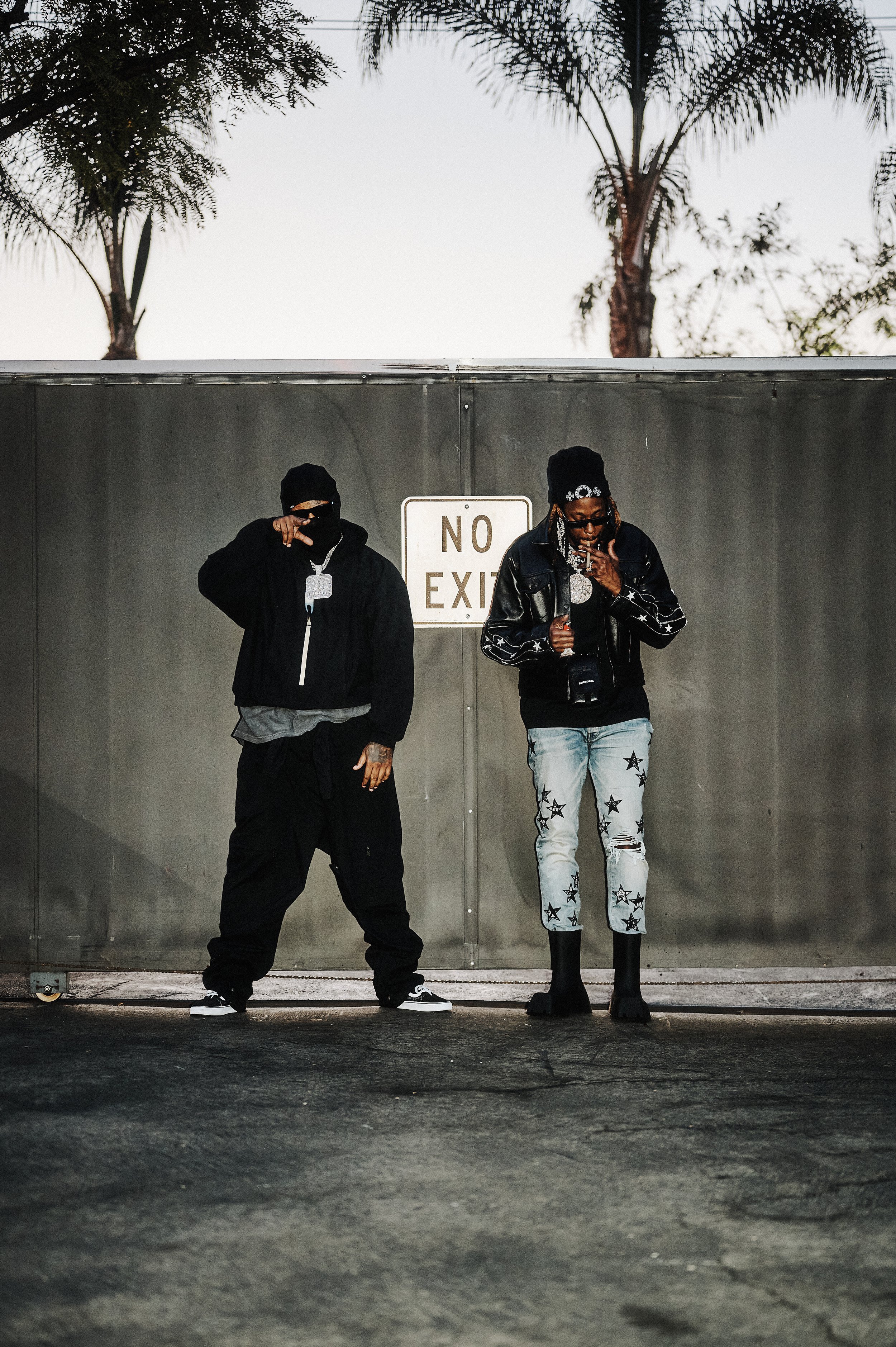 Two men in urban streetwear standing in front of a metal fence with a 'No Exit' sign, palm trees are visible in the background.