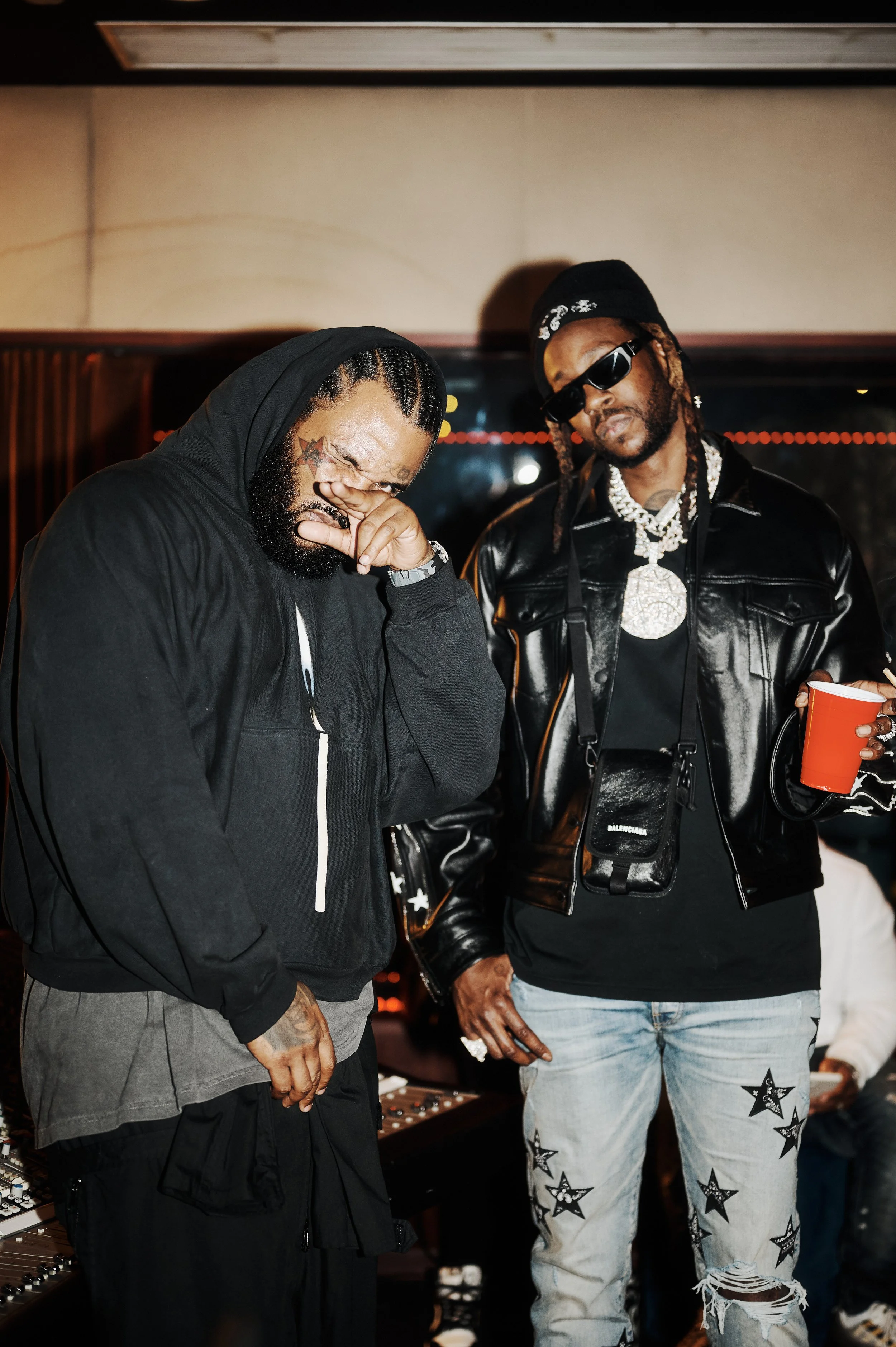 Two men in a music studio, one with braids and sunglasses wearing a black leather jacket and graphic jeans, the other with tattoos and dreadlocks wiping his eye, dressed in a dark hoodie and gray shirt, with musical equipment in the background.