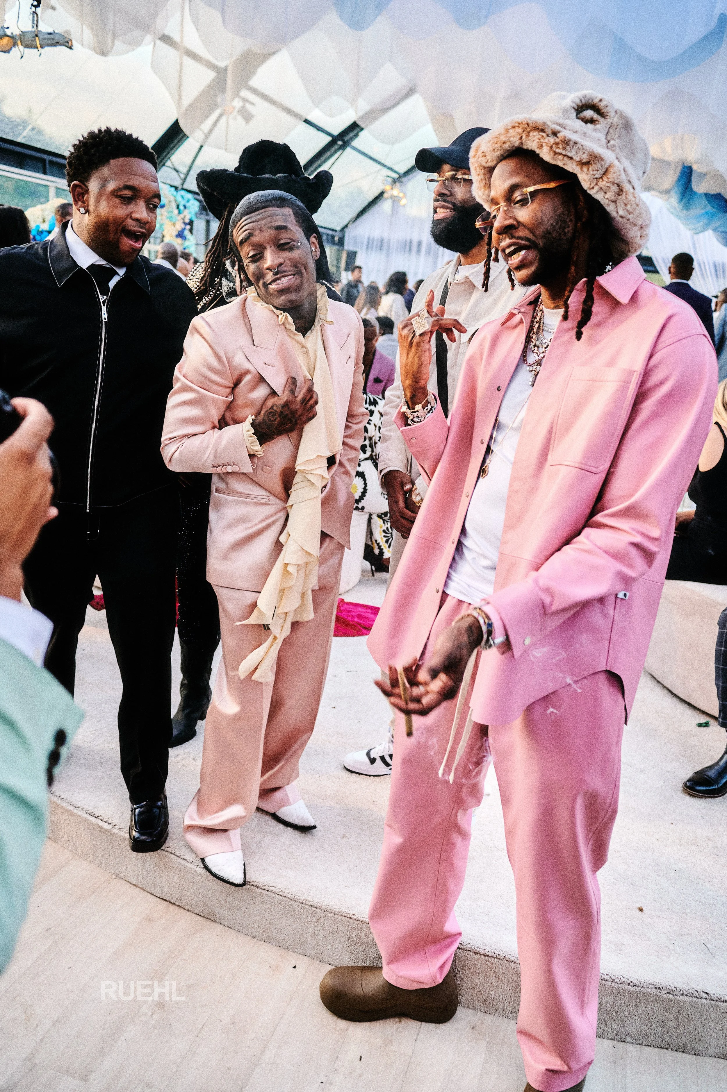 Group of men dressed in fashionable clothing, some in pink suits, engaged in conversation at an indoor event with a tent-like ceiling and other attendees in the background.