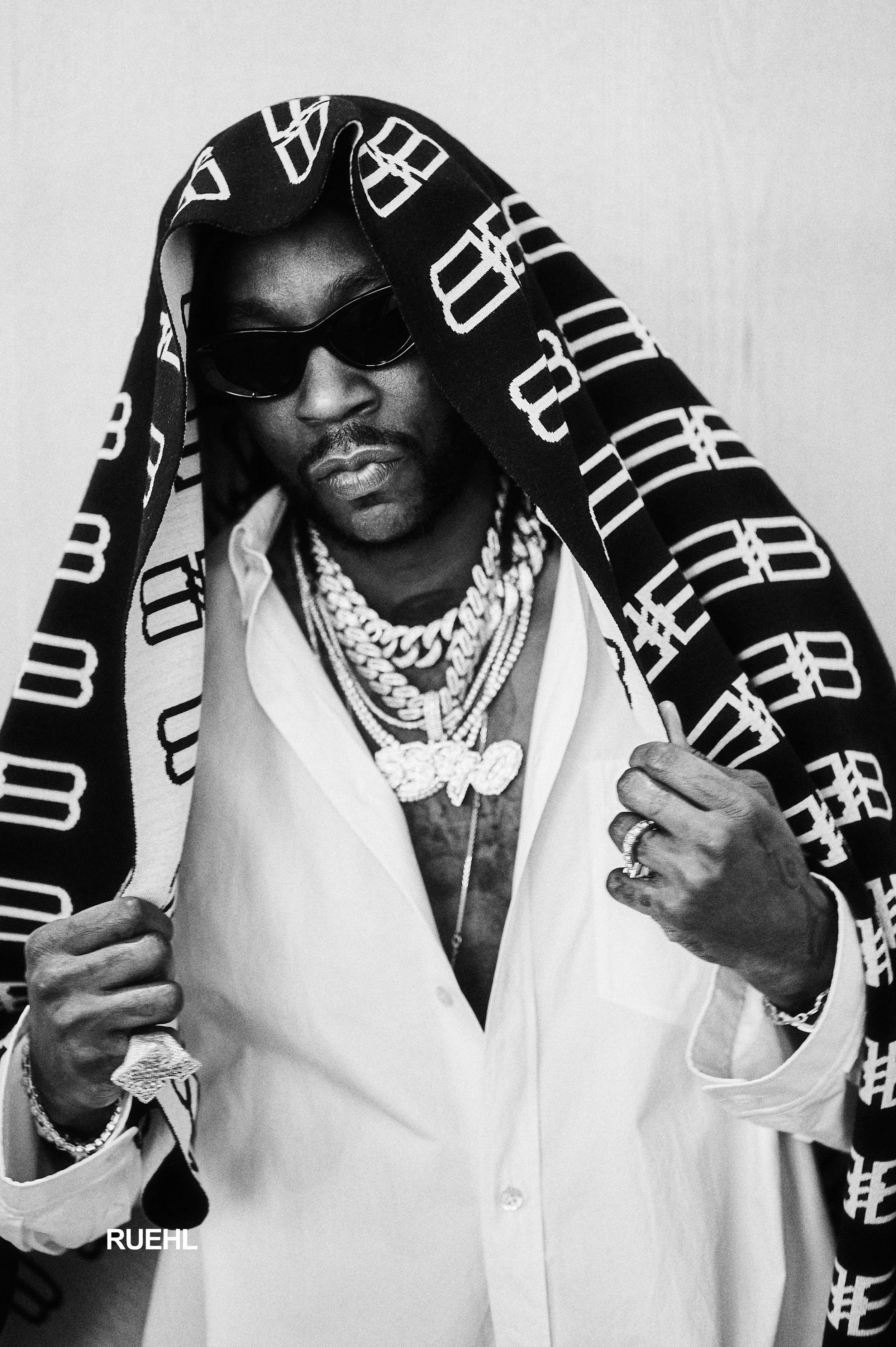 Black and white photo of a man wearing sunglasses, multiple layered chains, and rings, holding a cloth with a pattern over his head.