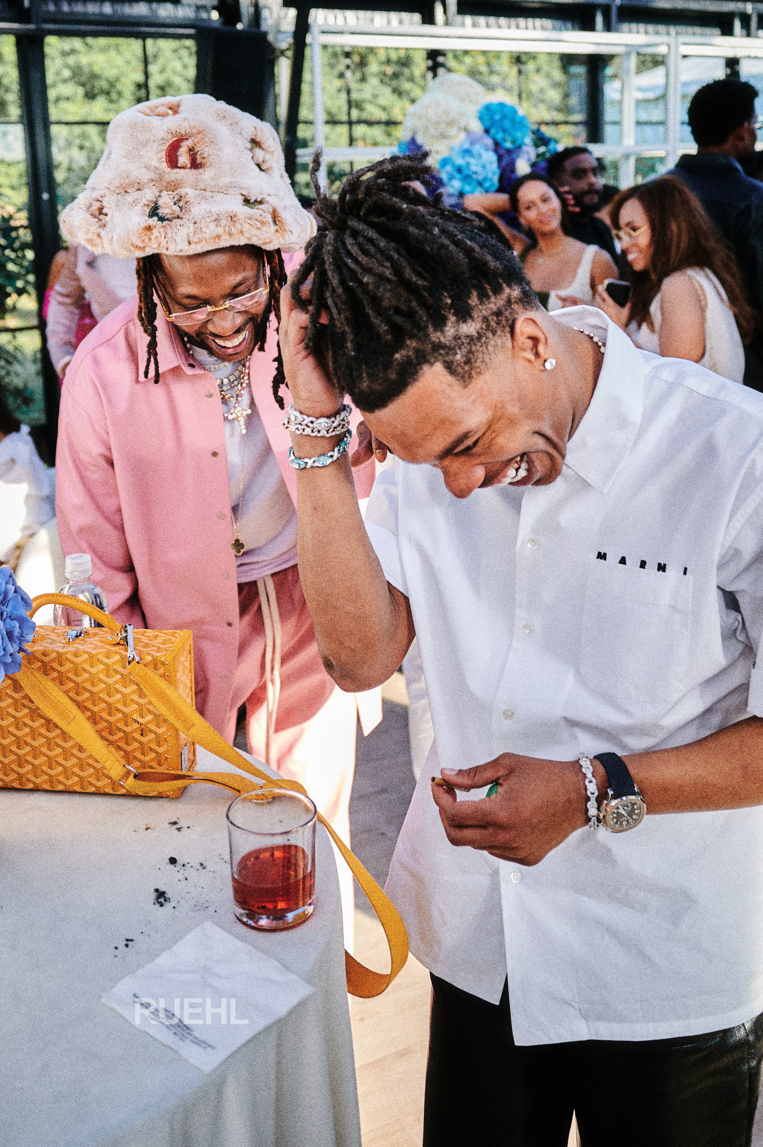 Two men laughing and enjoying themselves at a social gathering, one wearing a pink outfit and a bucket hat, the other in a white shirt with a logo, surrounded by other people in the background.