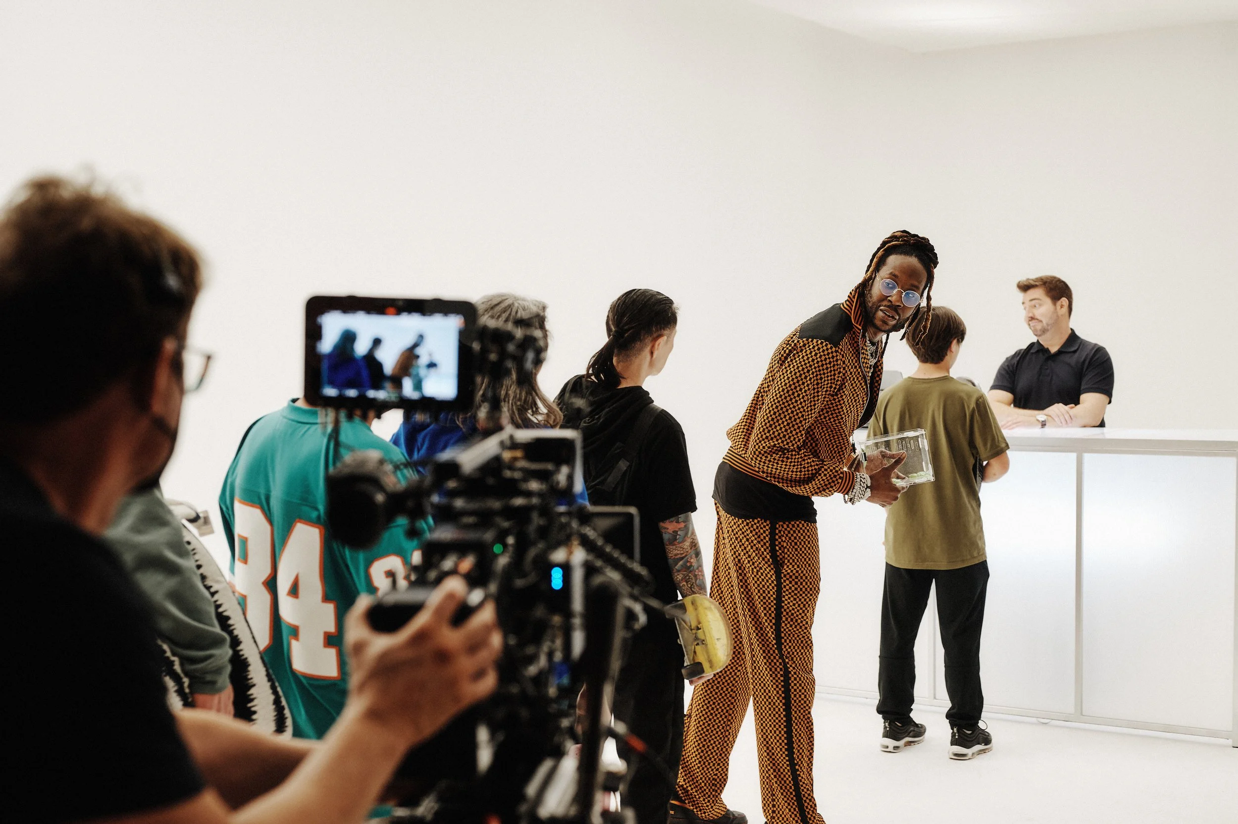 An art gallery or auction house with a group of people, including a man with dreadlocks in checkered clothing holding a glass container, standing in front of a white wall. Some people are observing or talking, and a camera operator is filming the sce