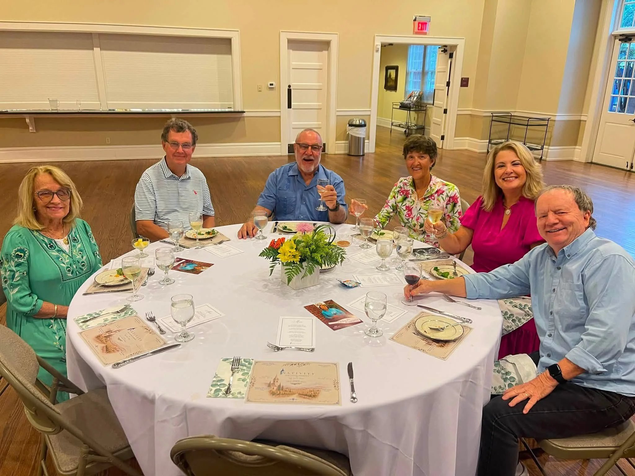 Six people sitting around a round dining table with a floral centerpiece and drinks, in a well-lit banquet hall.