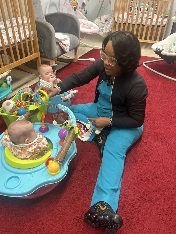 Woman playing with a baby in a colorful indoor play area with toys, cribs, and a red carpet.