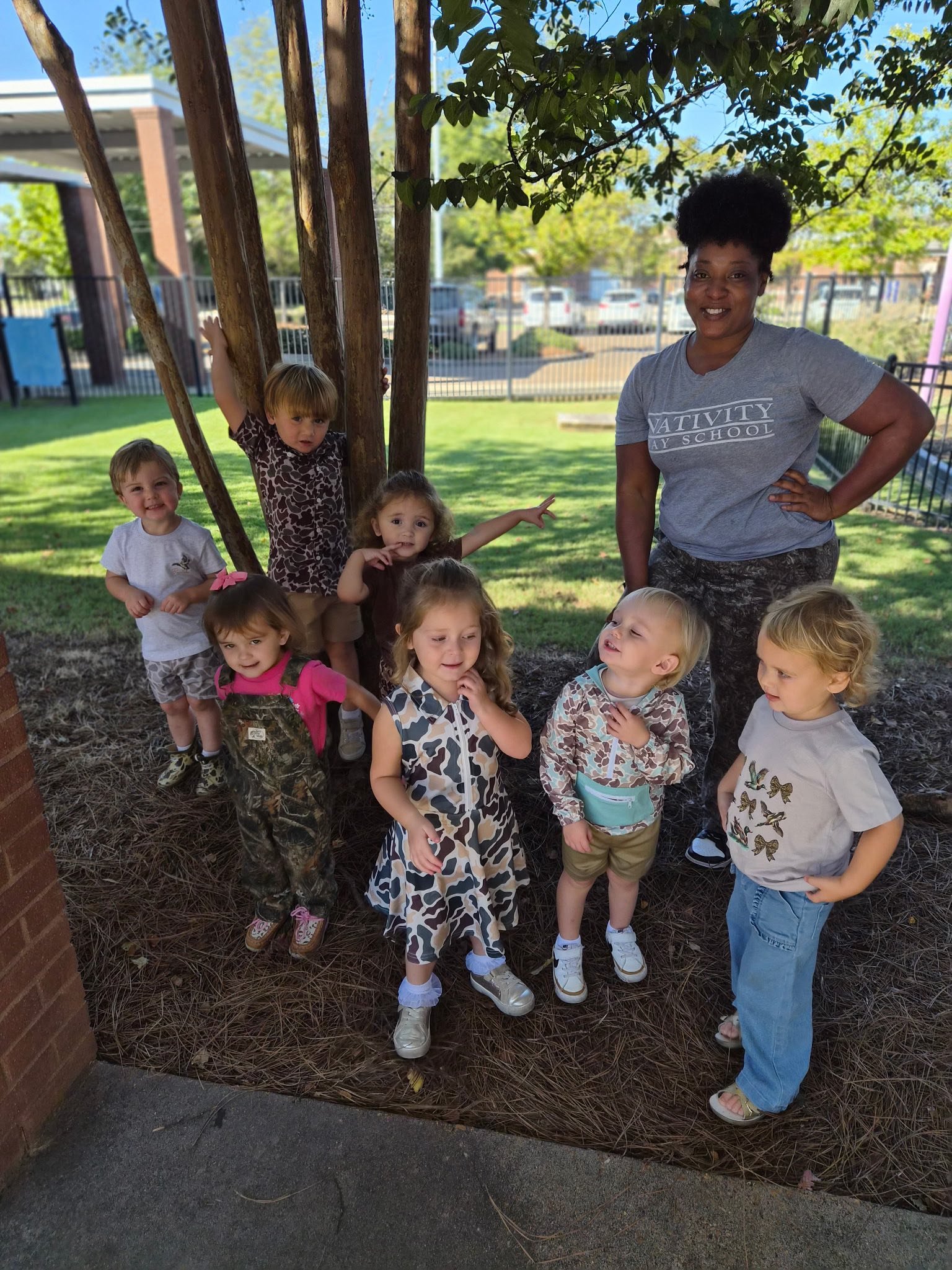 A group of young children and a woman outdoors in a backyard, some children are standing or sitting, and the woman is smiling with her hand on her hip, with trees, a fence, and parked cars in the background.