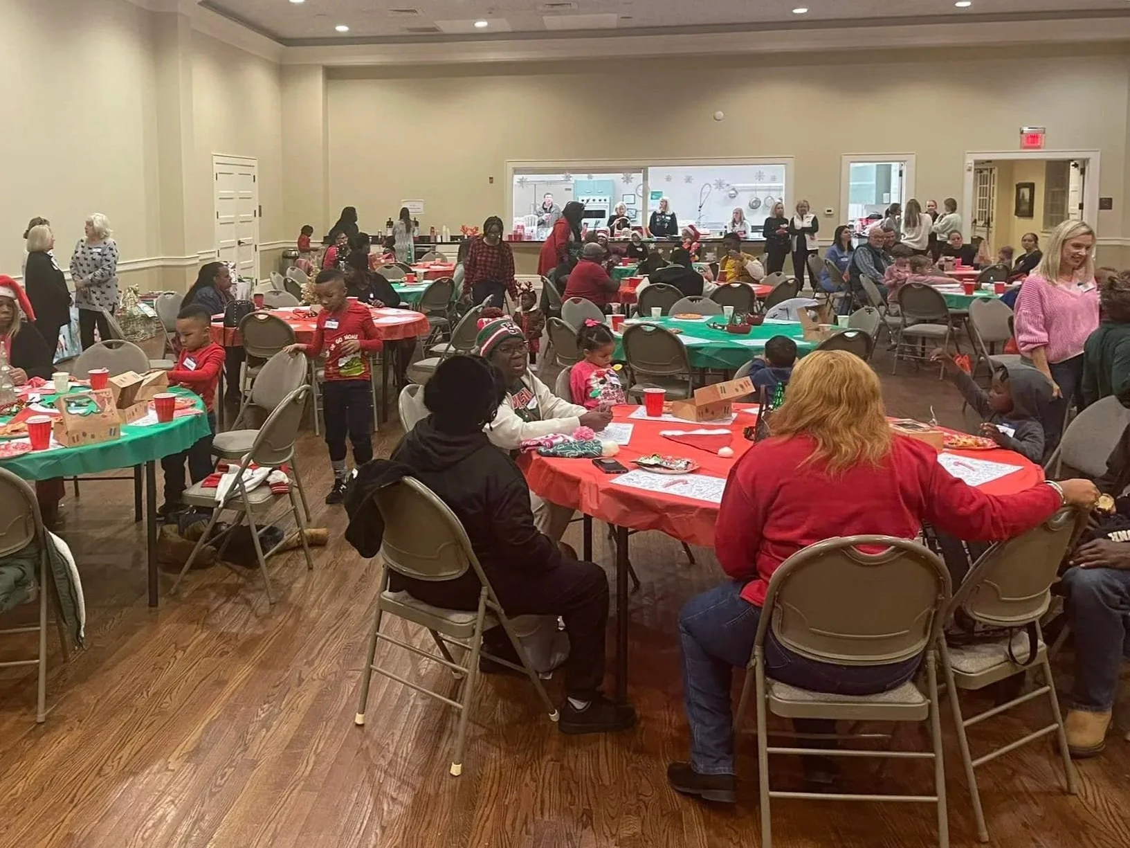 A large room filled with people sitting at round tables covered with red and green tablecloths. The tables have food, drinks, and gift boxes on them. There are children and adults, some standing and some seated, engaging in conversations and activities. In the background, a kitchen window shows people preparing food. The room has wooden floors and beige walls.