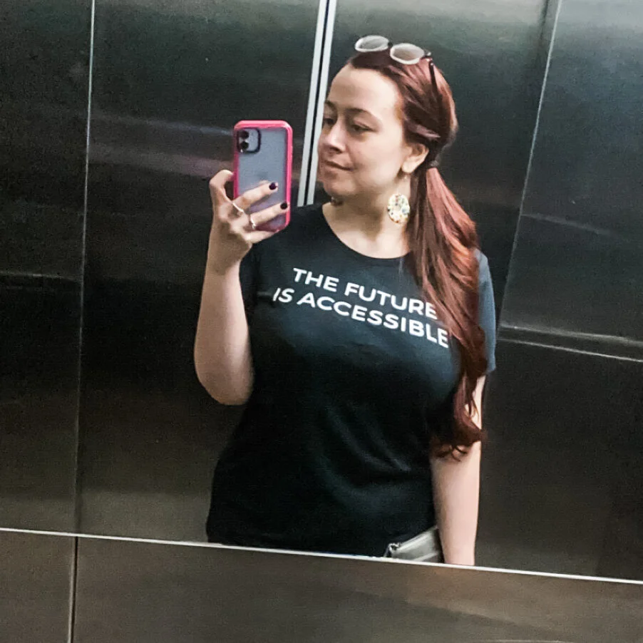Image Description: Maddie stands in an elevator and takes a seflie in the mirror. She is wearing a black t-shirt that says “The future is accessible” in white font. Her long red hair is in a side ponytail to the right and she holds a purple iphone i…
