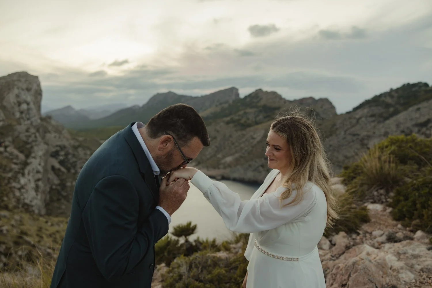 Groom kissing bride’s hand during their emotional elopement at Cap de Formentor, Mallorca. Surrounded by dramatic cliffs and sea views, a quiet and intimate wedding moment full of tenderness.
