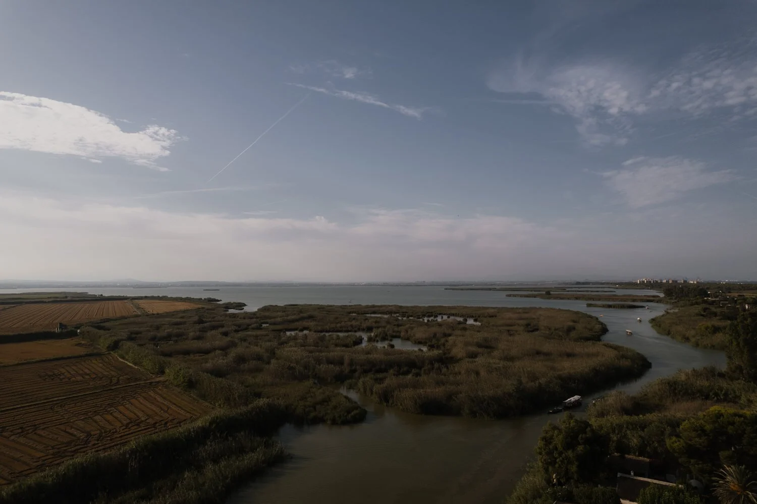 Wide aerial view of Albufera wetlands with winding waterways, rice fields, and boats under a soft afternoon sky near Valencia.