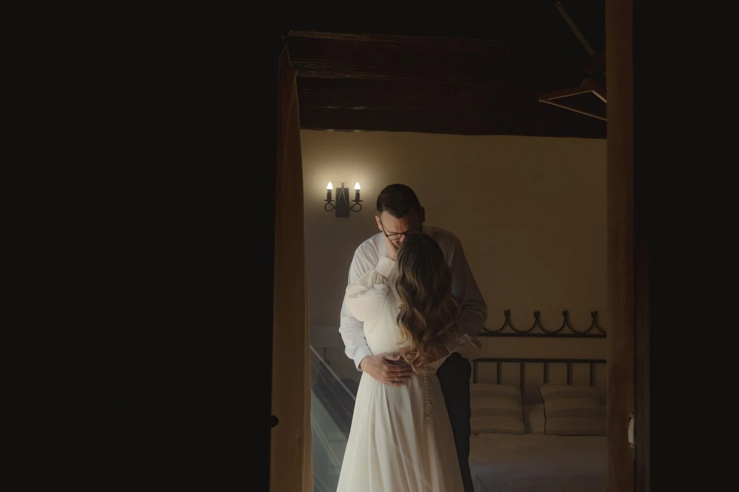 Emotional first look moment between bride and groom in a cozy rustic bedroom in Mallorca. The couple shares an intimate embrace, framed by soft light and wooden beams, captured by luxury elopement photographer Adrian Vilanova.