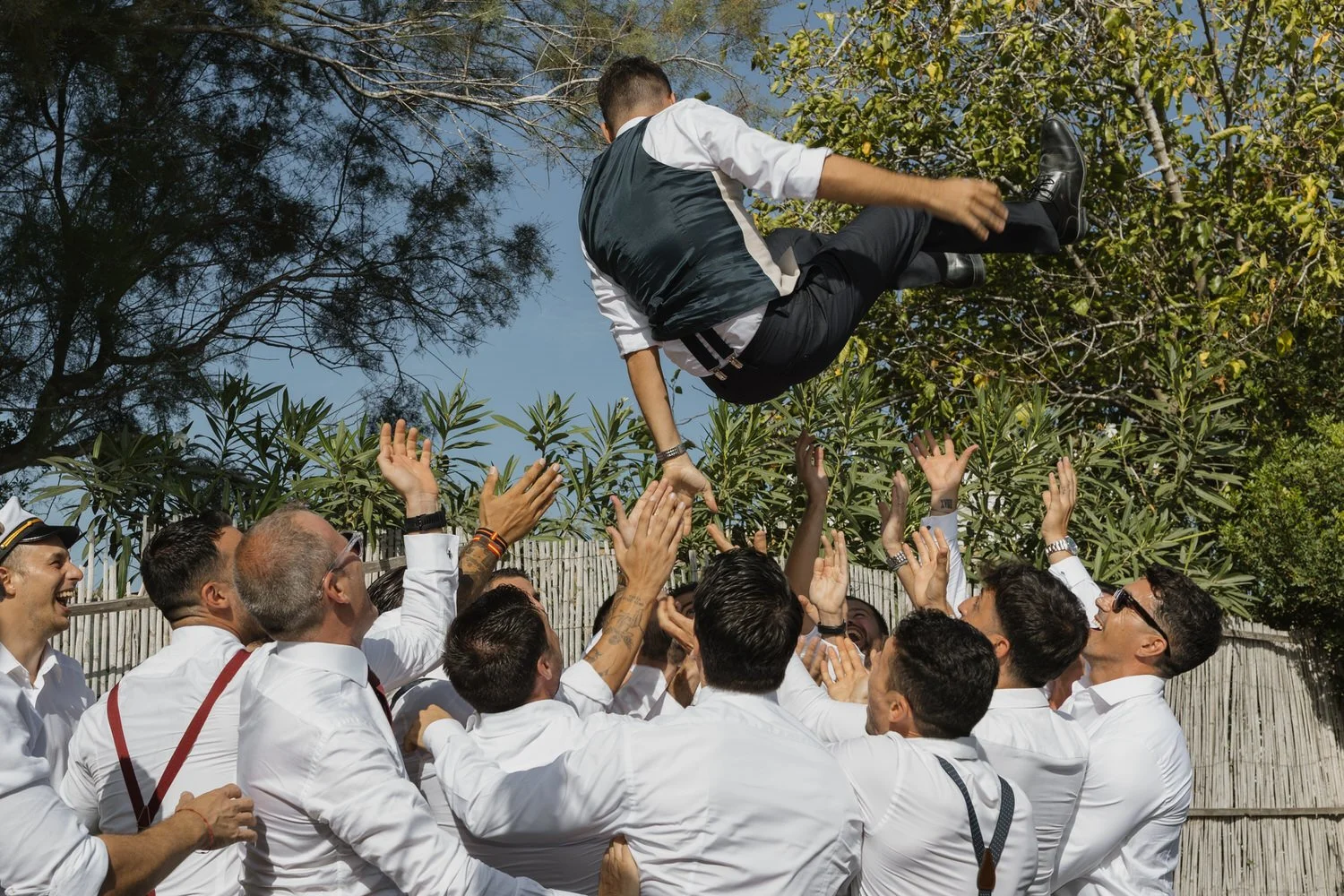 Groom being lifted into the air by friends during a joyful outdoor wedding celebration, captured mid-motion under a clear sky.