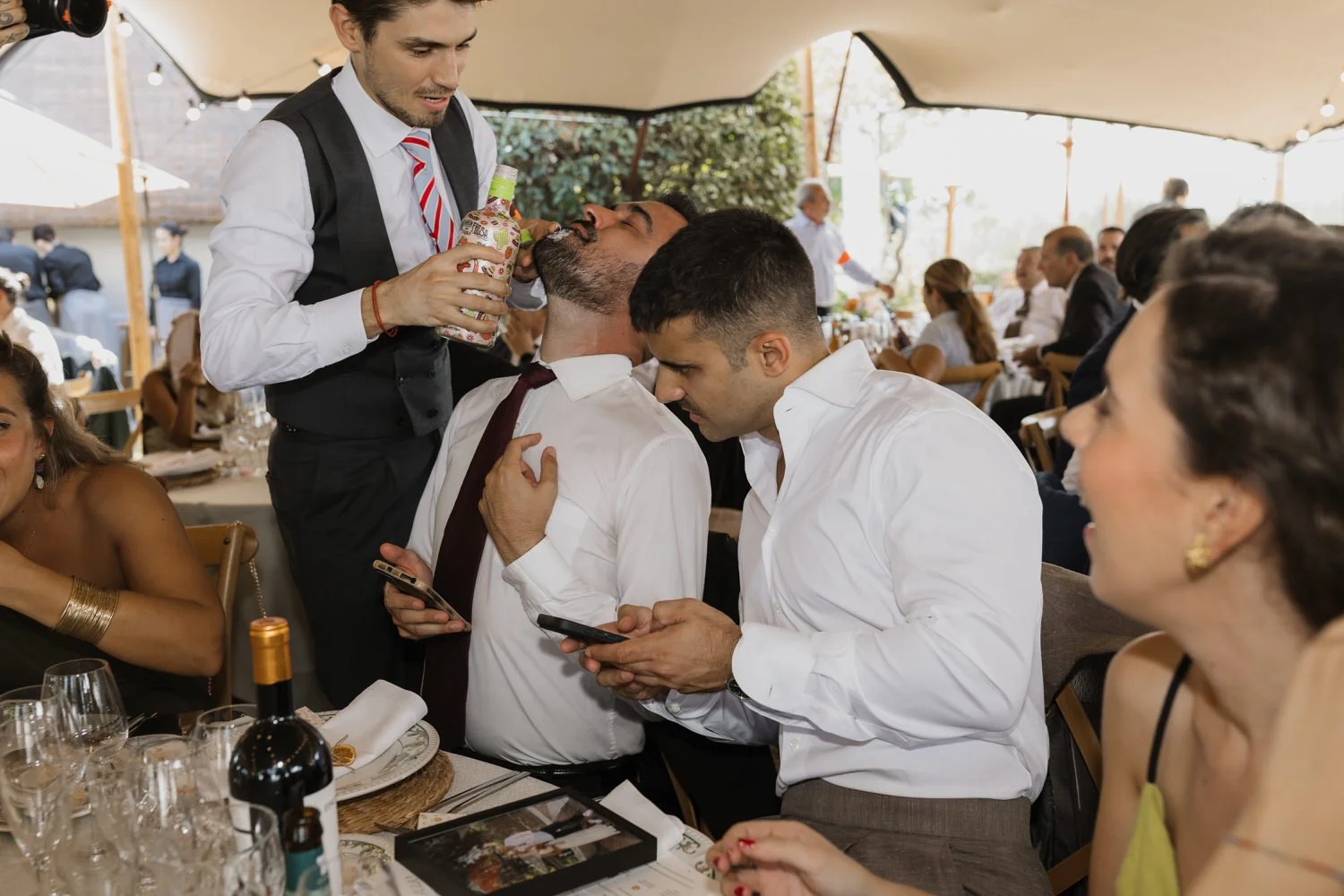 Friends laughing as one guest pours a drink into another’s mouth at a long wedding table during an energetic outdoor reception.