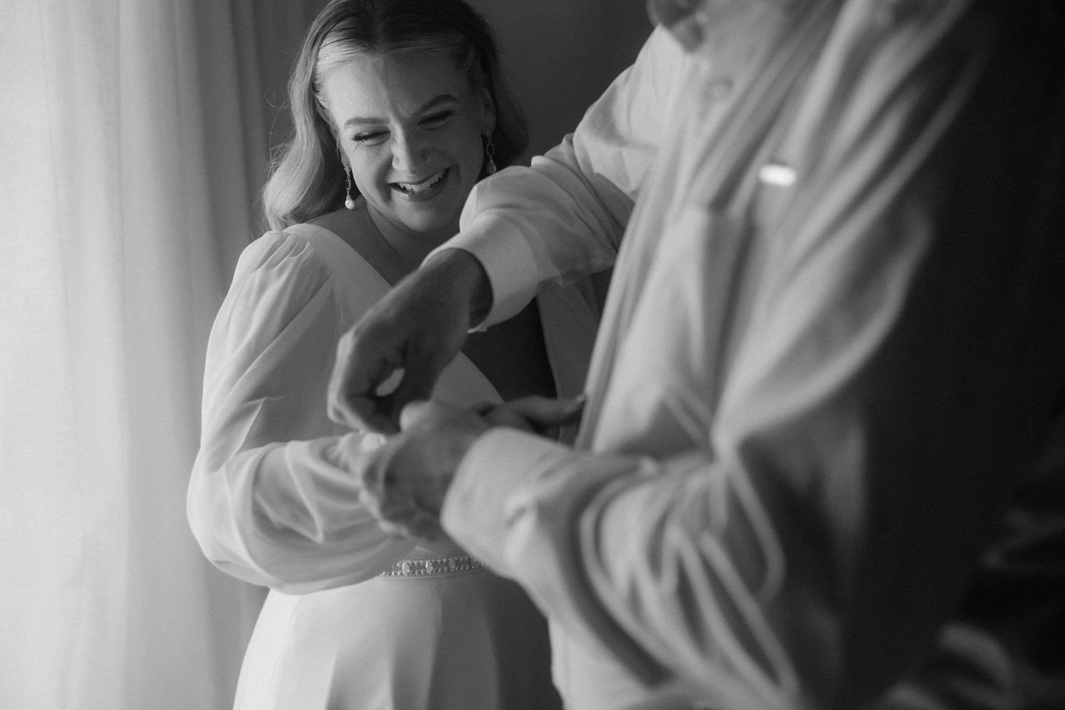 Bride smiling with joy as her partner helps her adjust her sleeve before their intimate elopement ceremony in Mallorca. A candid black and white moment captured by Adrian Vilanova, destination wedding photographer.