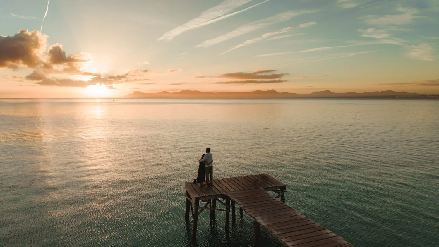 Couple standing on a wooden pier at sunrise in Mallorca, sharing a quiet moment by the sea during their elopement.