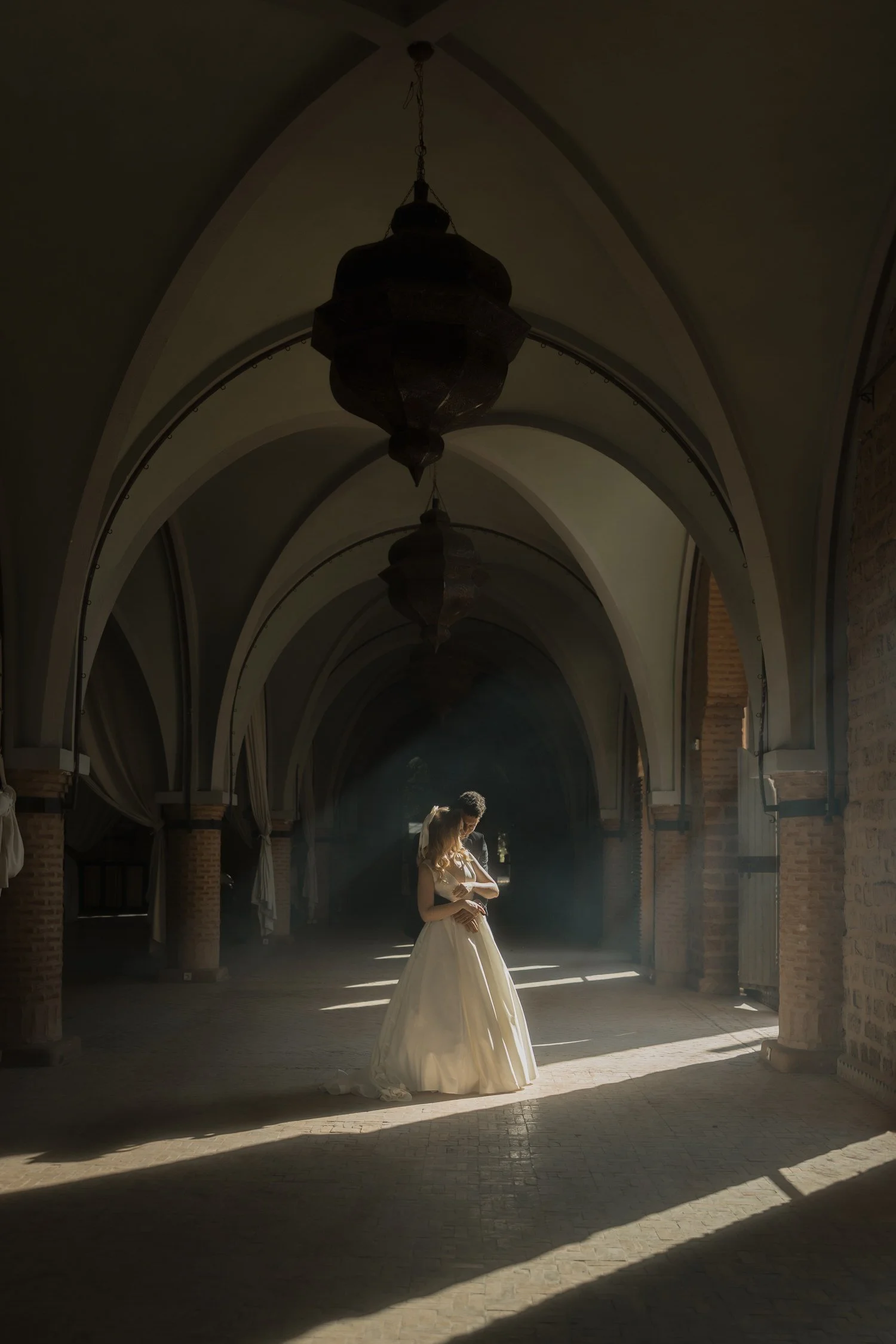 Bride and groom embracing under the arched halls of Beldi Country Club, Marrakech, with soft morning light and dust particles creating a cinematic atmosphere.