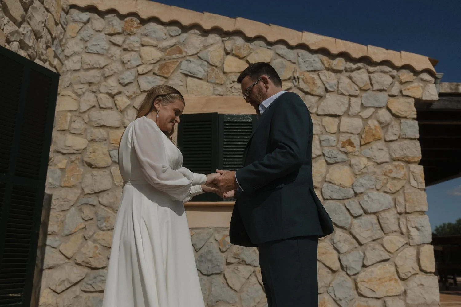 The couple stands in front of a rustic stone wall, holding hands and sharing an emotional, heartfelt moment under the Mediterranean sun.