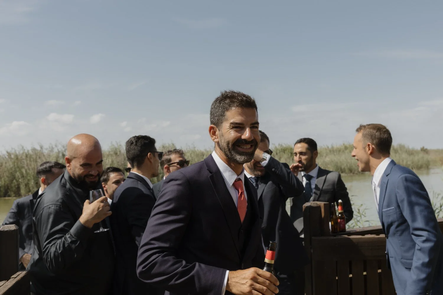 Guests sharing drinks and laughter by the water during an outdoor wedding celebration, with reeds and blue sky in the background.