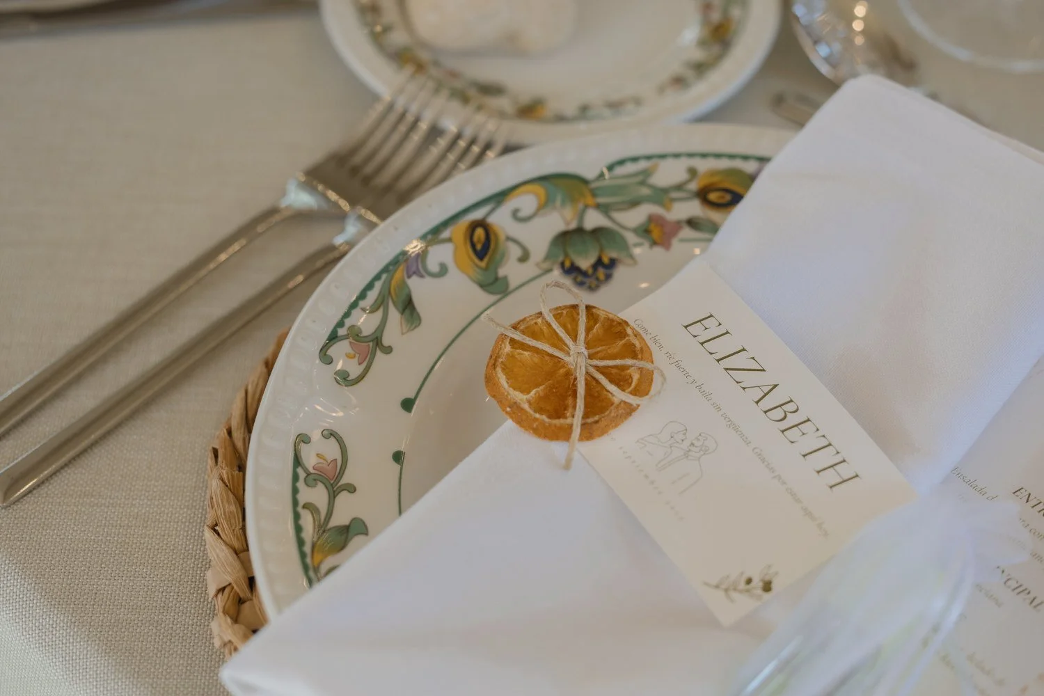 Elegant wedding table setting with hand-painted ceramic plates, white linen napkin, and a personalized place card tied with a dried orange slice.
