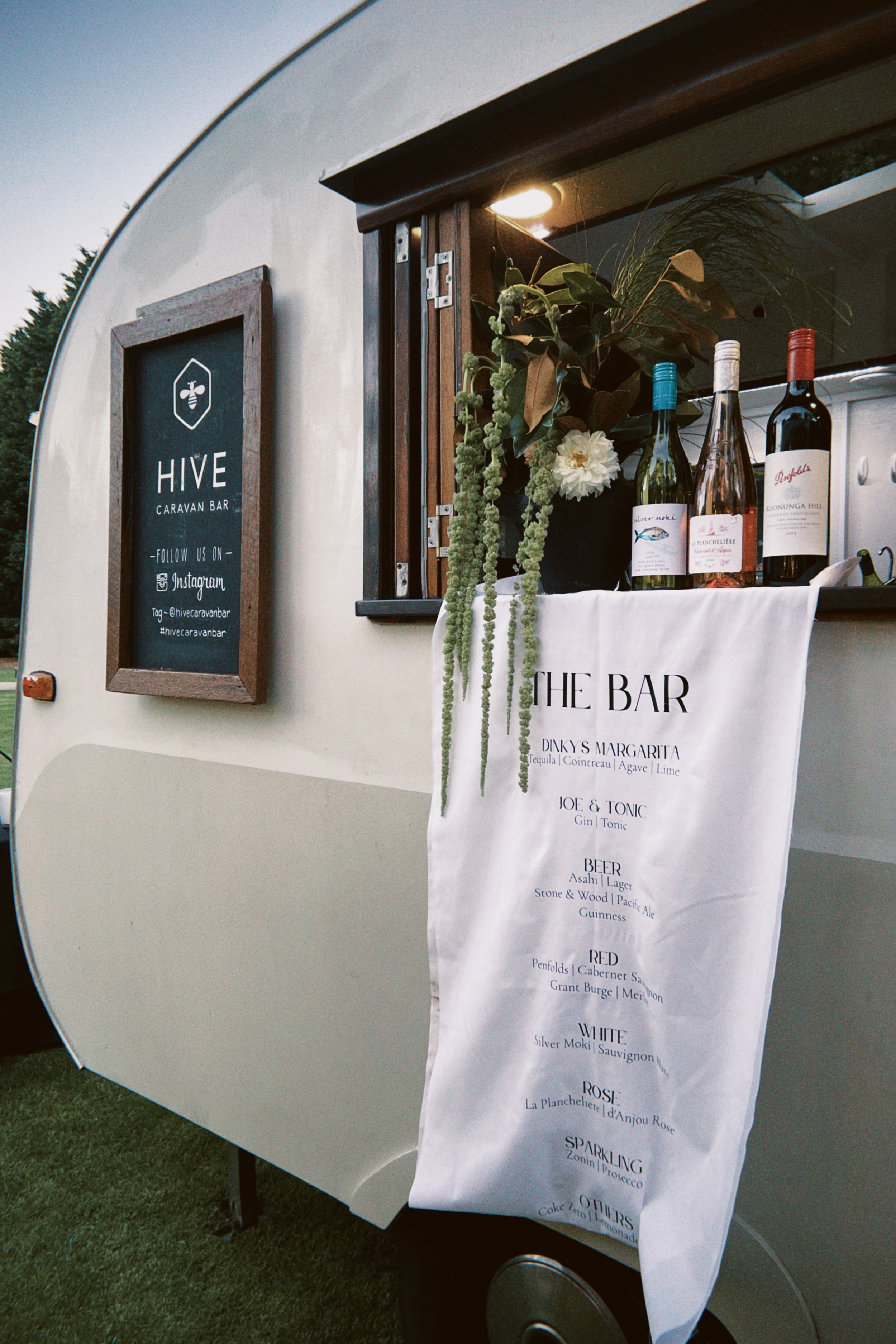 Exterior of hive caravan bar with a sign that reads 'HIVE Caravan Bar' and a menu listing various alcoholic beverages including wine, beer, and cocktails, with decorative plants and bottles displayed.
