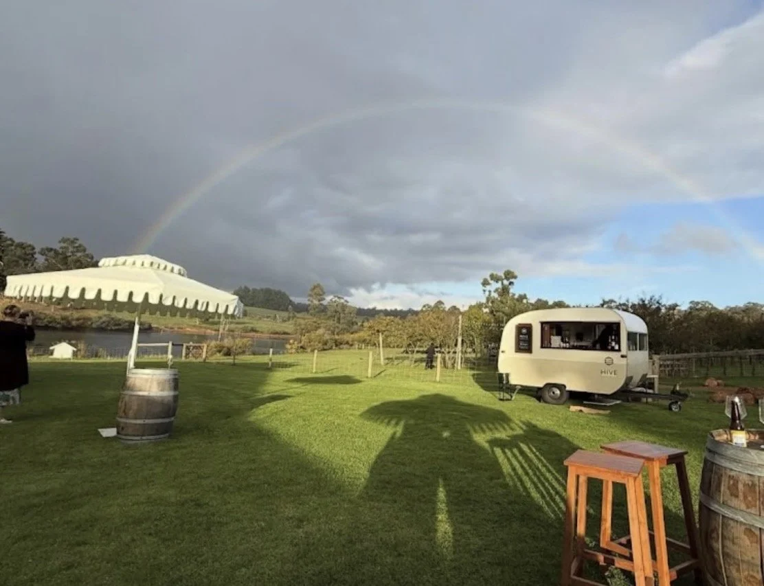 A scenic outdoor scene with green grass, a rainbow in the sky, the hive caravan bar, a barrel, a small table, a large umbrella, and some people in the distance.