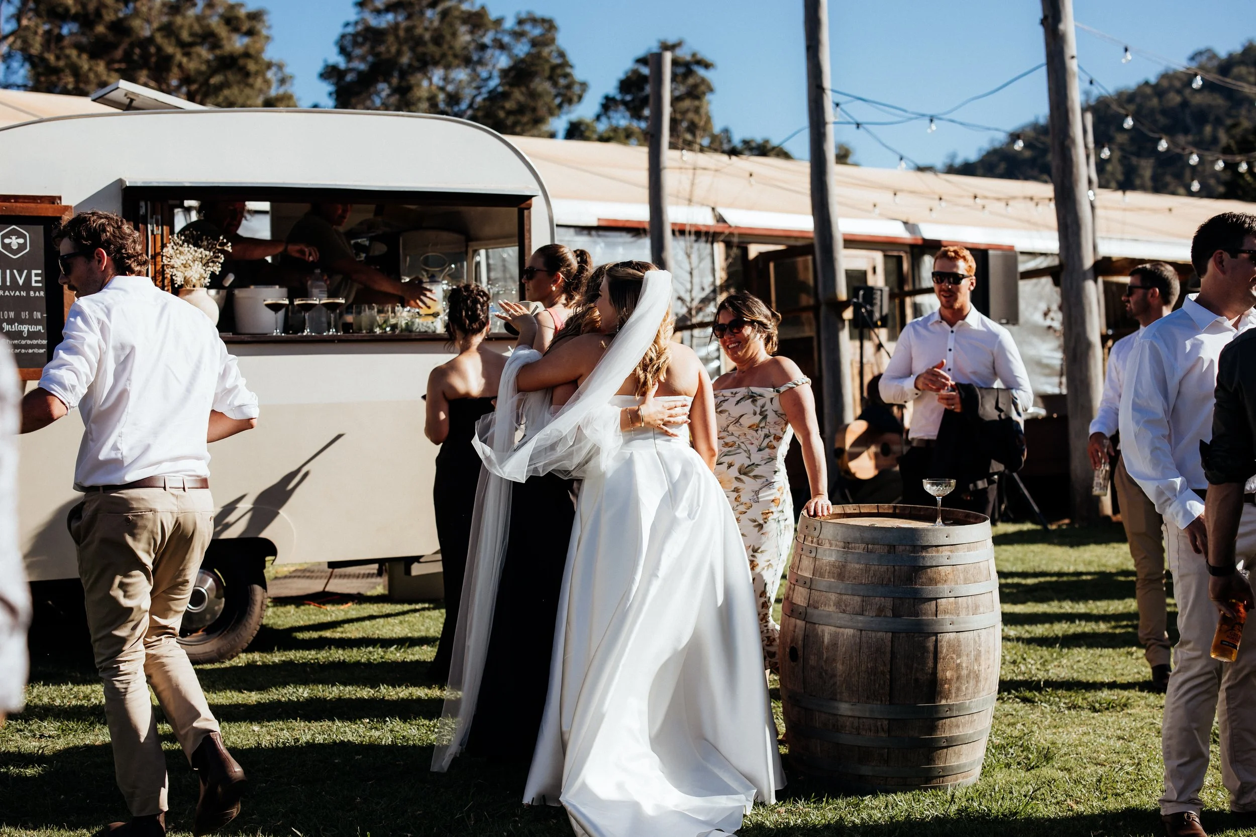 People dancing outdoors at a wedding reception, with a white hive caravan bar serving drinks in the background and a hillside covered with trees under a clear blue sky.