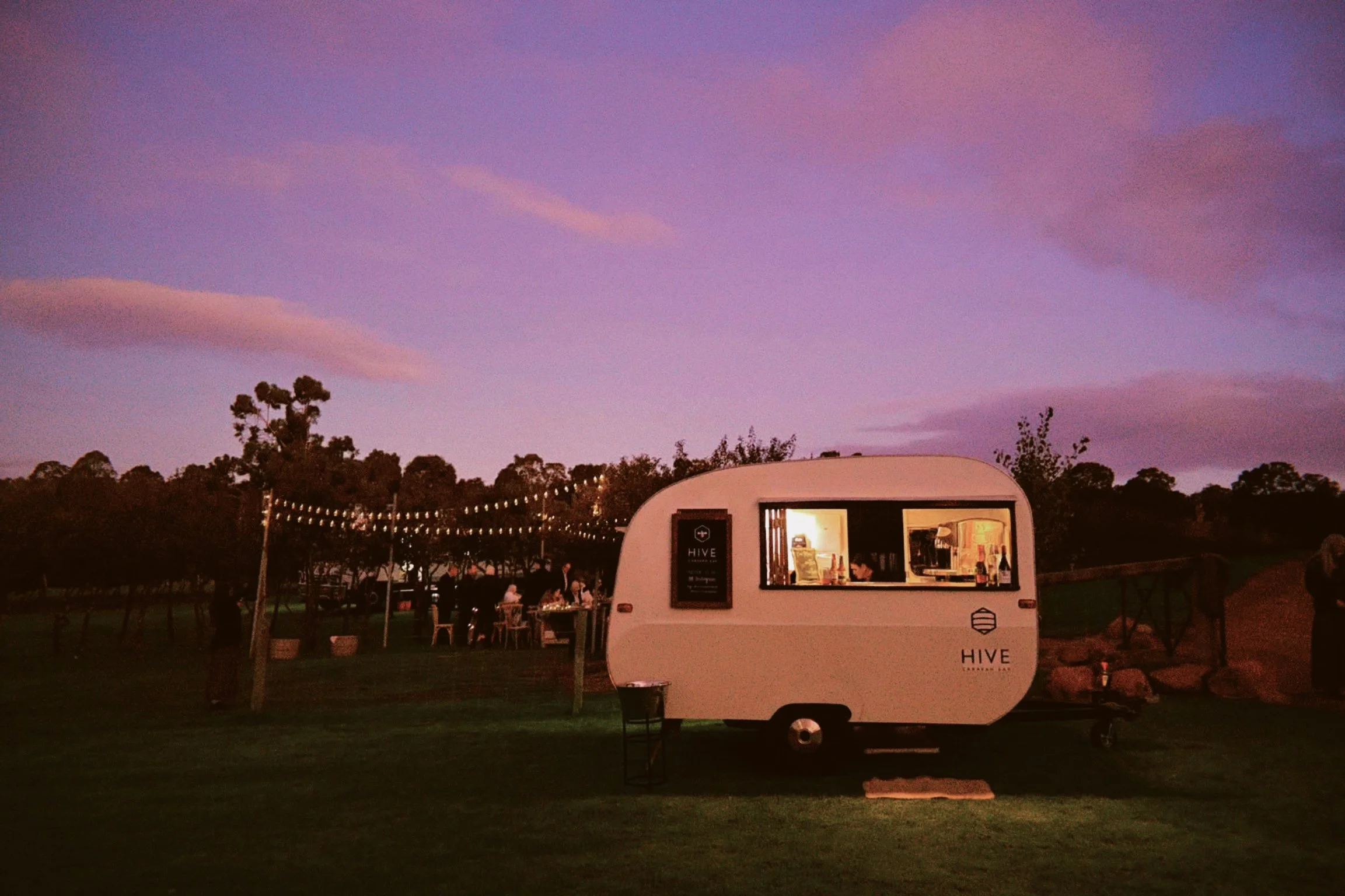 Twilight sky with pink and purple clouds over a park with trees, string lights, and a white caravan bar labeled 'HIVE' serving drinks/ beverages to south west customers.