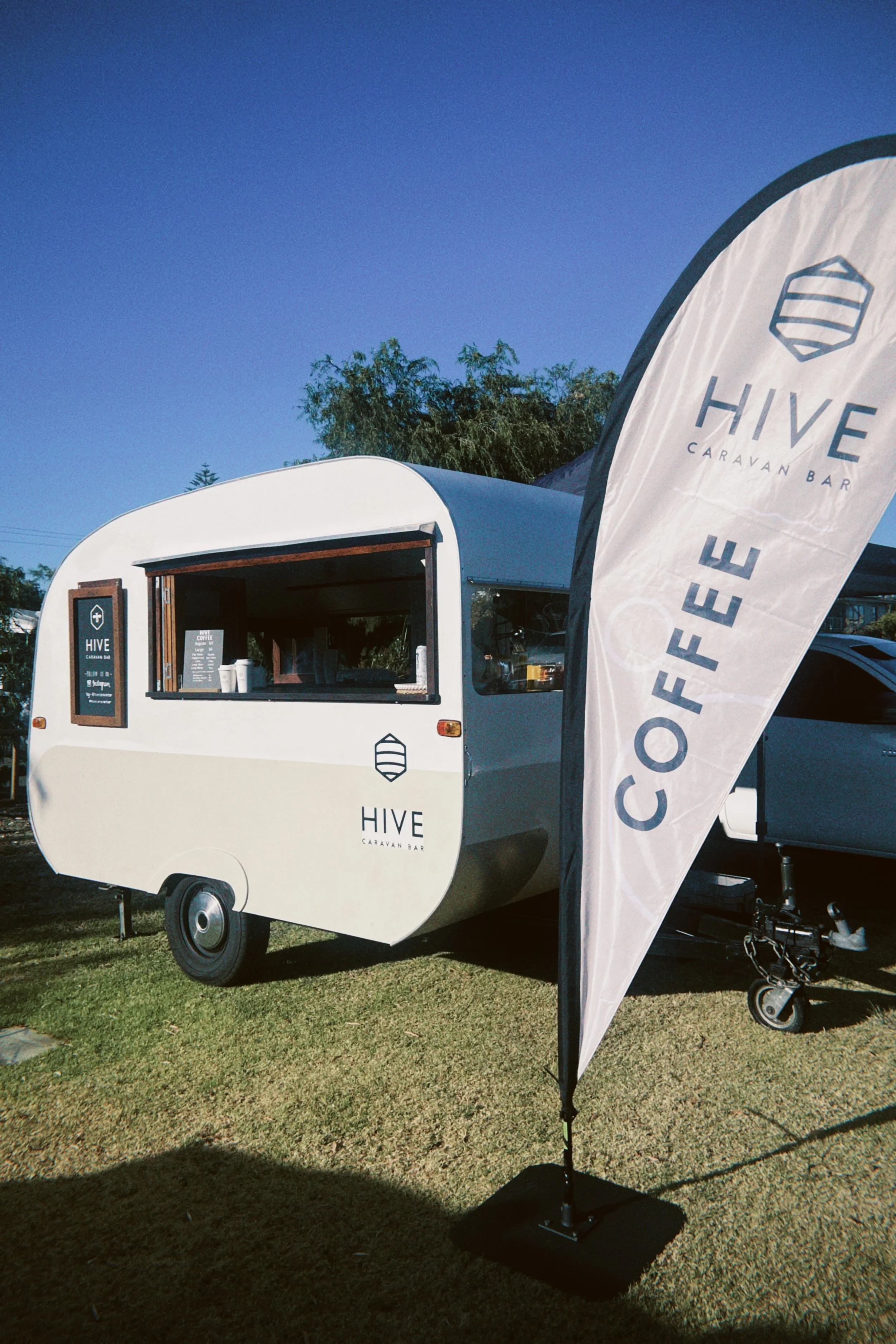 A white caravan with a serving window and a menu sign on the side, set up outdoors on grass. There is a tall flag with the logo and name "HIVE Caravan Bar" in front. Trees and a clear blue sky are in the background.