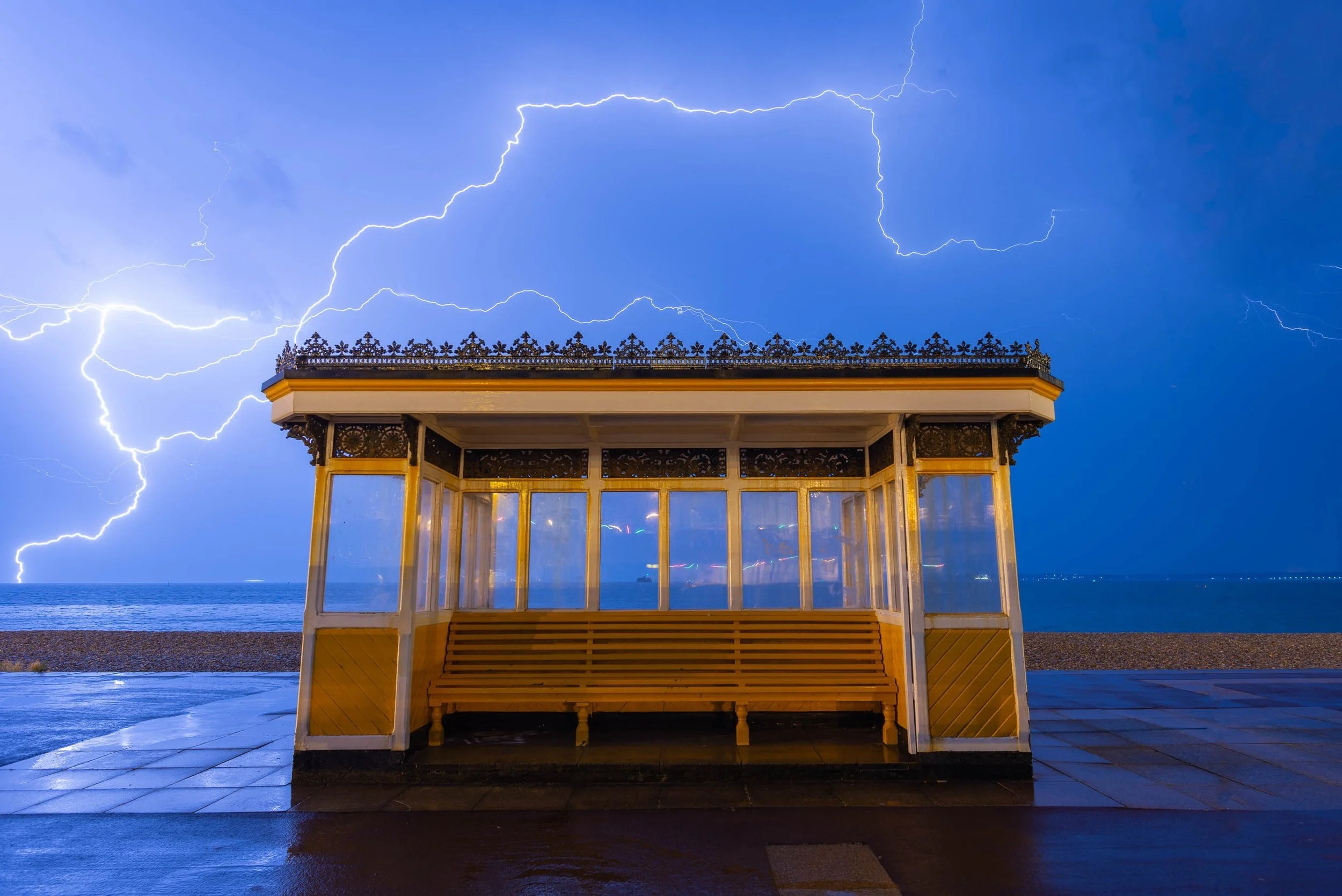 A wooden beach shelter with ornate details on its roof, situated on a wet promenade by the sea during a lightning storm, with visible bolts of lightning in the sky.
